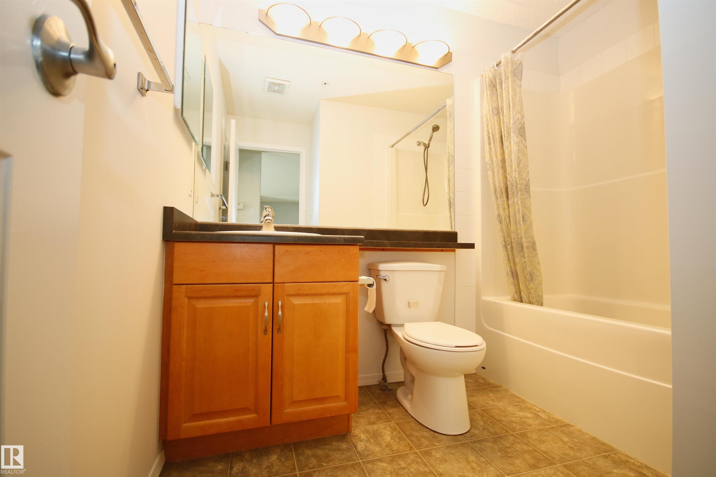 Full bathroom featuring a wood-finish vanity with dark countertop, an integrated sink, and a mirrored wall - 311 2098 Blackmud Creek Drive, Edmonton, AB