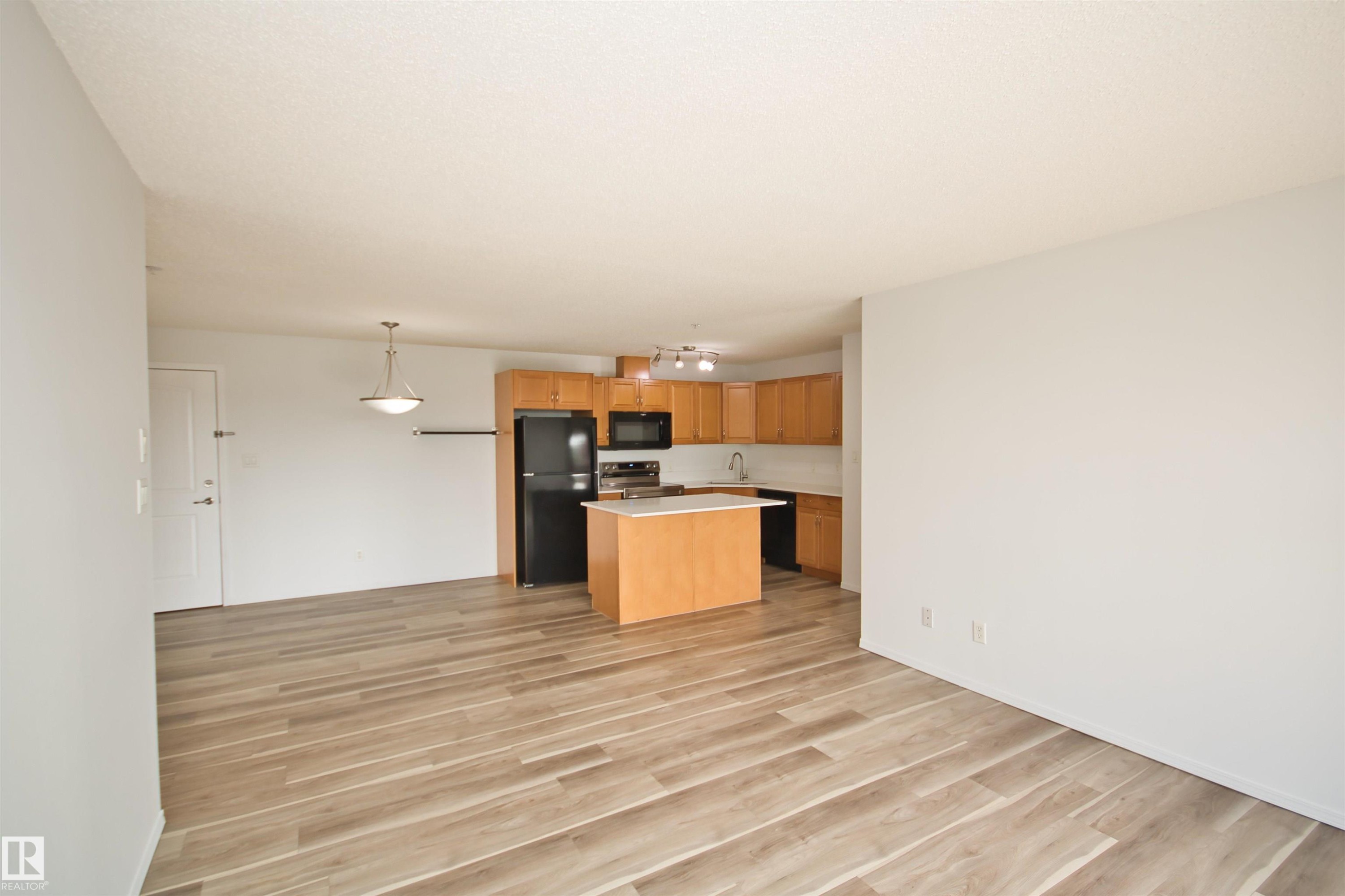 Open-concept living area featuring wood-finish flooring, a kitchen island, pendant lighting, built-in cabinetry, and black appliances - 311 2098 Blackmud Creek Drive, Edmonton, AB