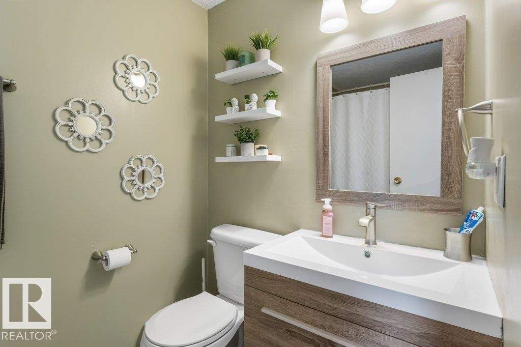 Bathroom featuring a wood-finish vanity with an integrated white sink, brushed nickel faucet, framed mirror, and floating wall shelves - 171 Ridgewood, St. Albert, AB