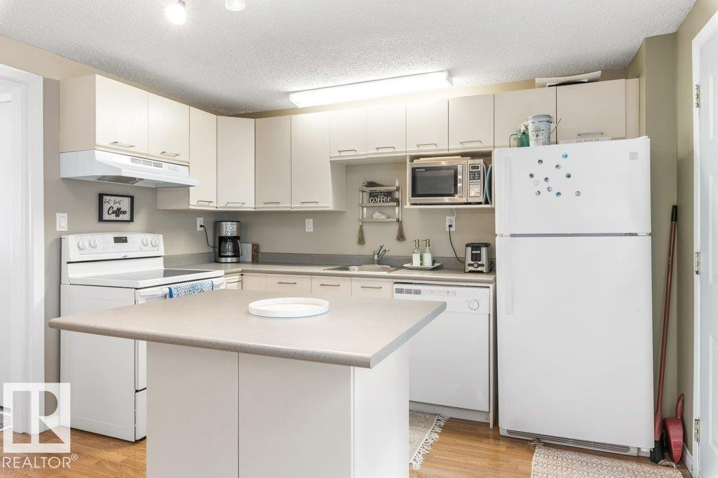 Kitchen featuring a central island with a solid surface countertop, white cabinetry with silver-tone hardware, wood-finish flooring, and white appliances including a range, refrigerator, and dishwasher - 171 Ridgewood, St. Albert, AB