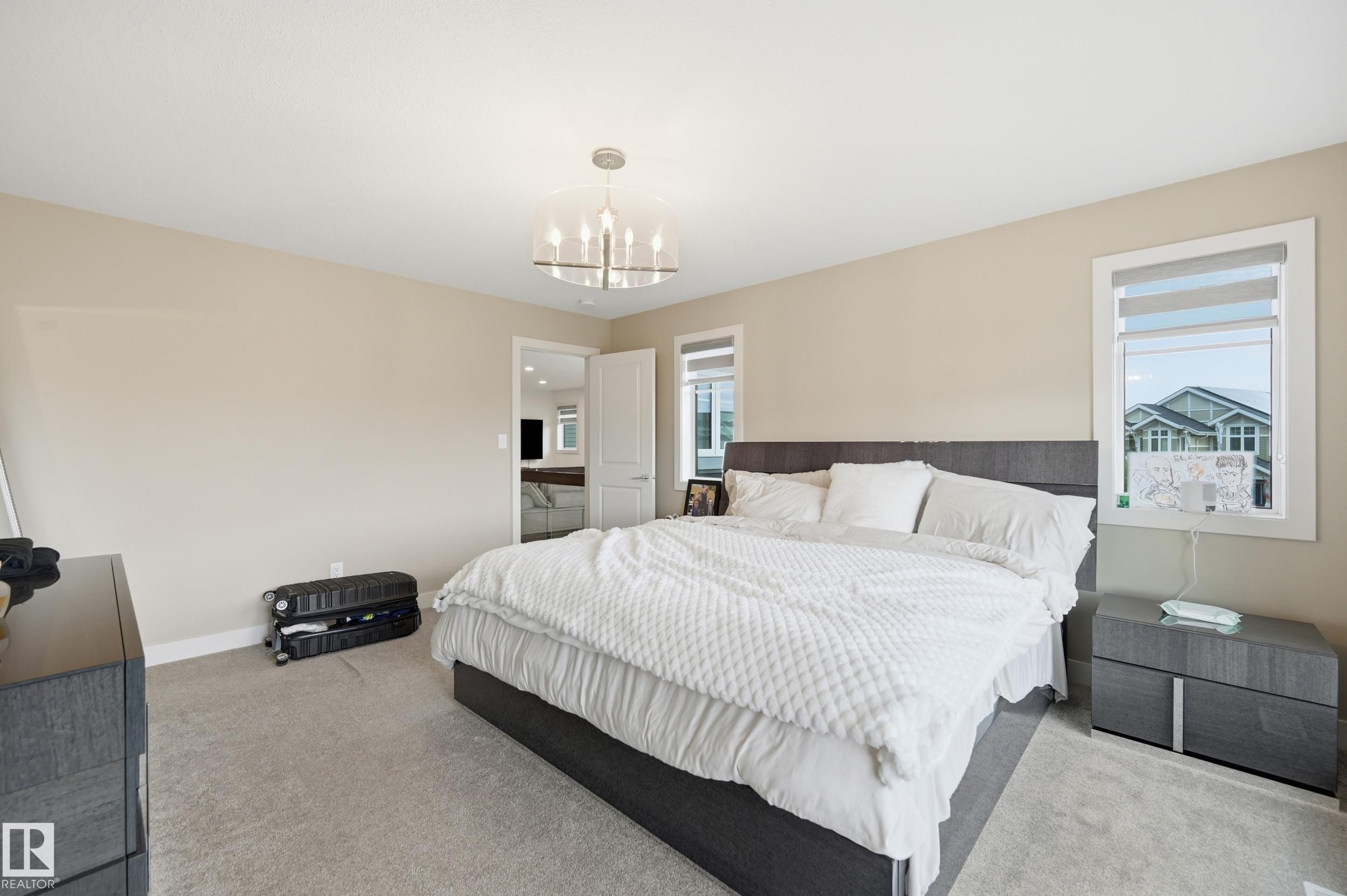 Spacious bedroom featuring light neutral wall tones, plush carpet flooring, a contemporary chandelier, and multiple windows with roller blinds - 76 Jacobs Close, St. Albert, AB