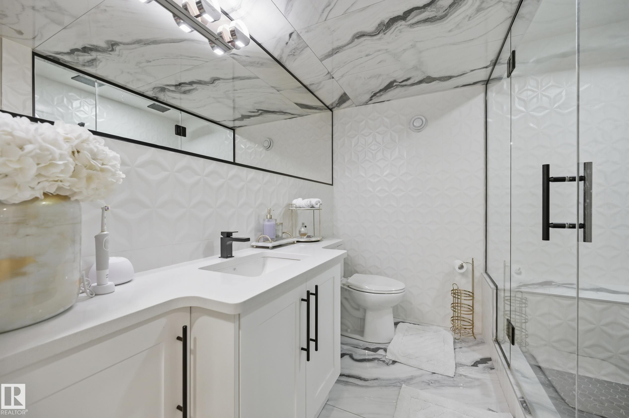 Bathroom featuring a white vanity with integrated sink and matte black faucet, wall-mounted mirror, geometric accent wall tiles, and a glass-enclosed shower - 76 Jacobs Close, St. Albert, AB