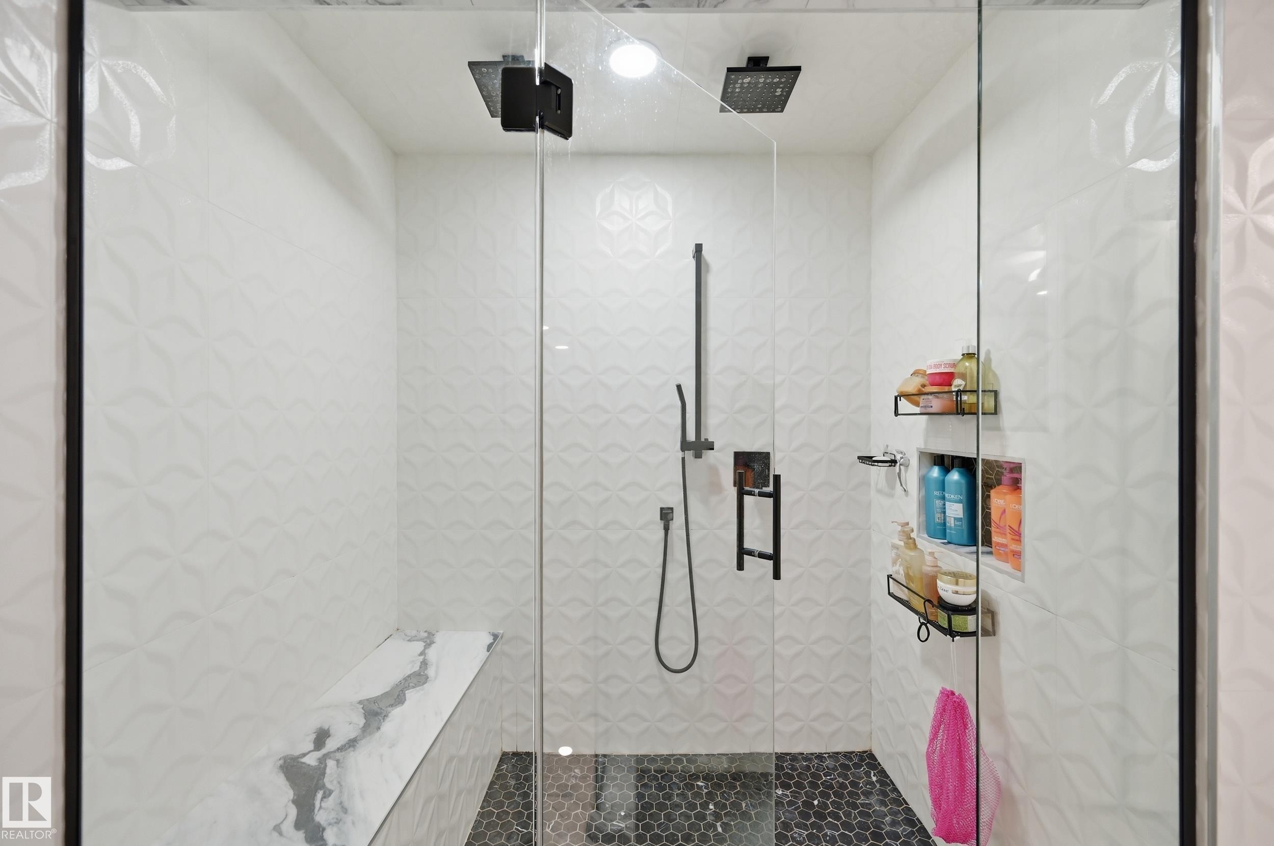 Glass-enclosed shower featuring dual matte black rain shower heads, a wall-mounted hand shower, a built-in bench with a stone-finish surface, patterned white tile walls, and a black hex tile floor - 76 Jacobs Close, St. Albert, AB