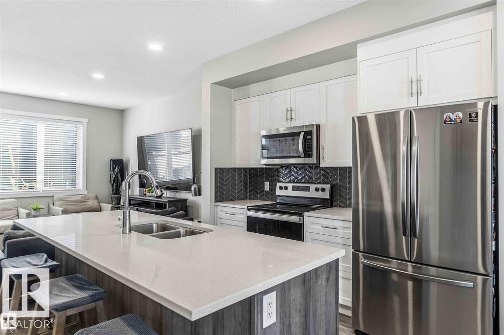 Open-concept kitchen featuring a large island with an undermount double-basin sink, stainless steel appliances, white cabinetry, and a chevron pattern tile backsplash - 1908 25 Street Nw, Edmonton, AB