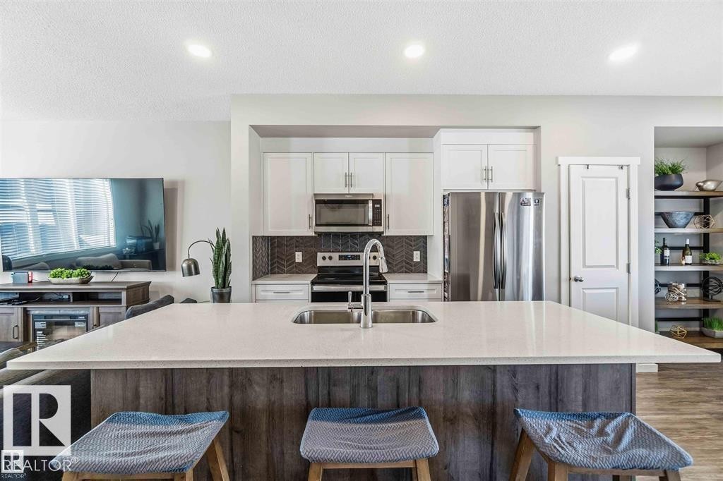 Kitchen featuring a large central island with an undermount sink, white cabinetry, stainless steel appliances, and a herringbone-patterned tile backsplash - 1908 25 Street Nw, Edmonton, AB