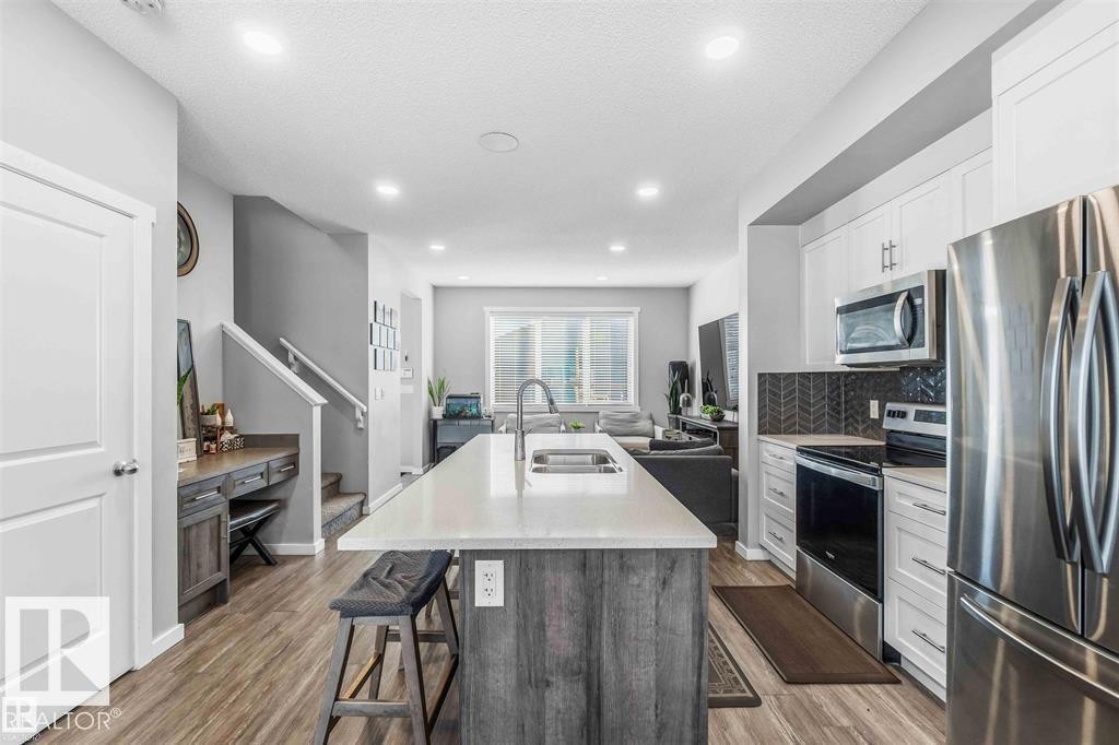 Contemporary kitchen featuring an oversized island with a built-in sink, stainless steel appliances, white cabinetry, and a dark herringbone tile backsplash - 1908 25 Street Nw, Edmonton, AB