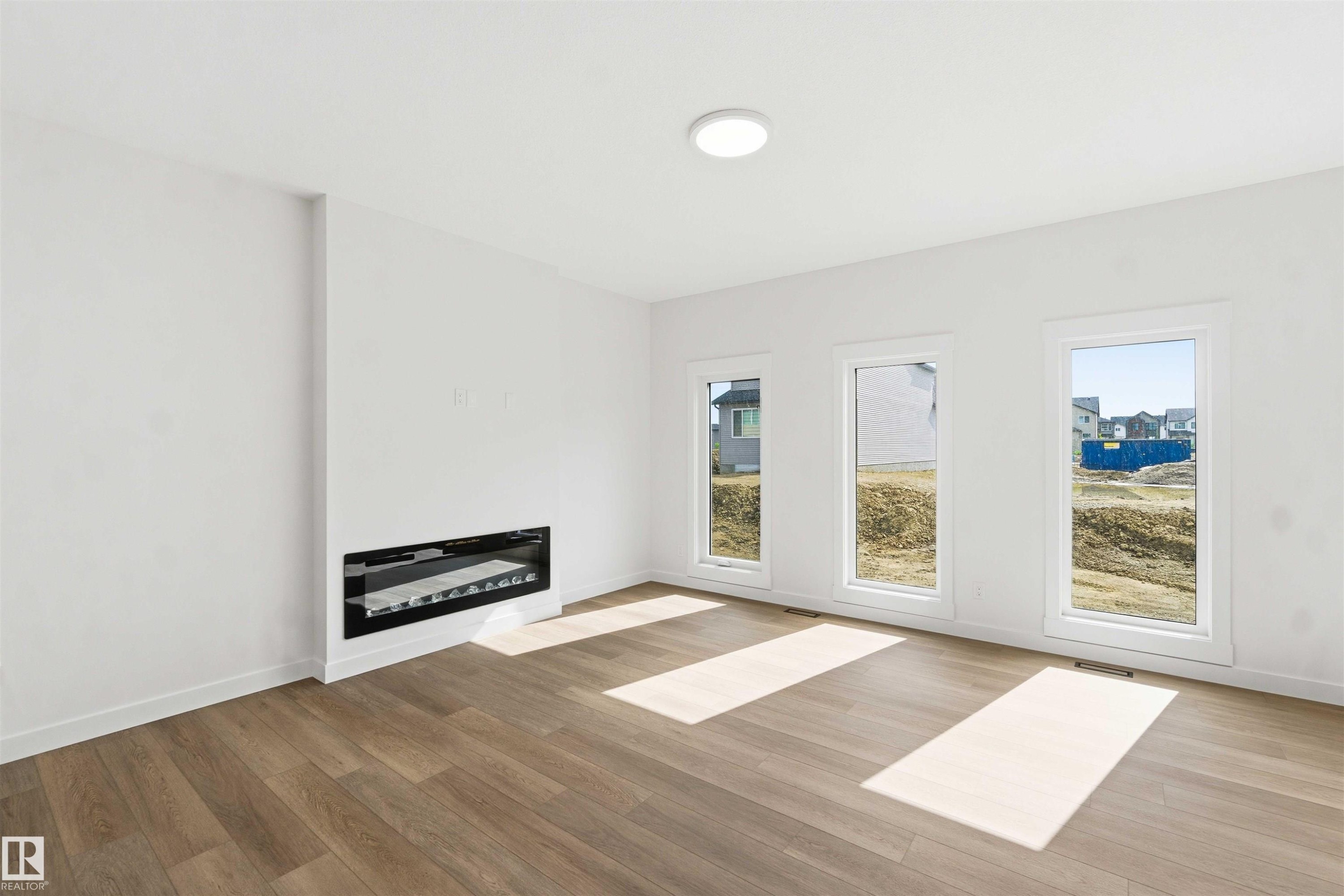 Open living area featuring wood-finish flooring, a linear built-in fireplace, three vertical windows, white trim, and a flush-mount ceiling light fixture - 1085 Cristall Crescent Sw, Edmonton, AB - Indoor With Fireplace