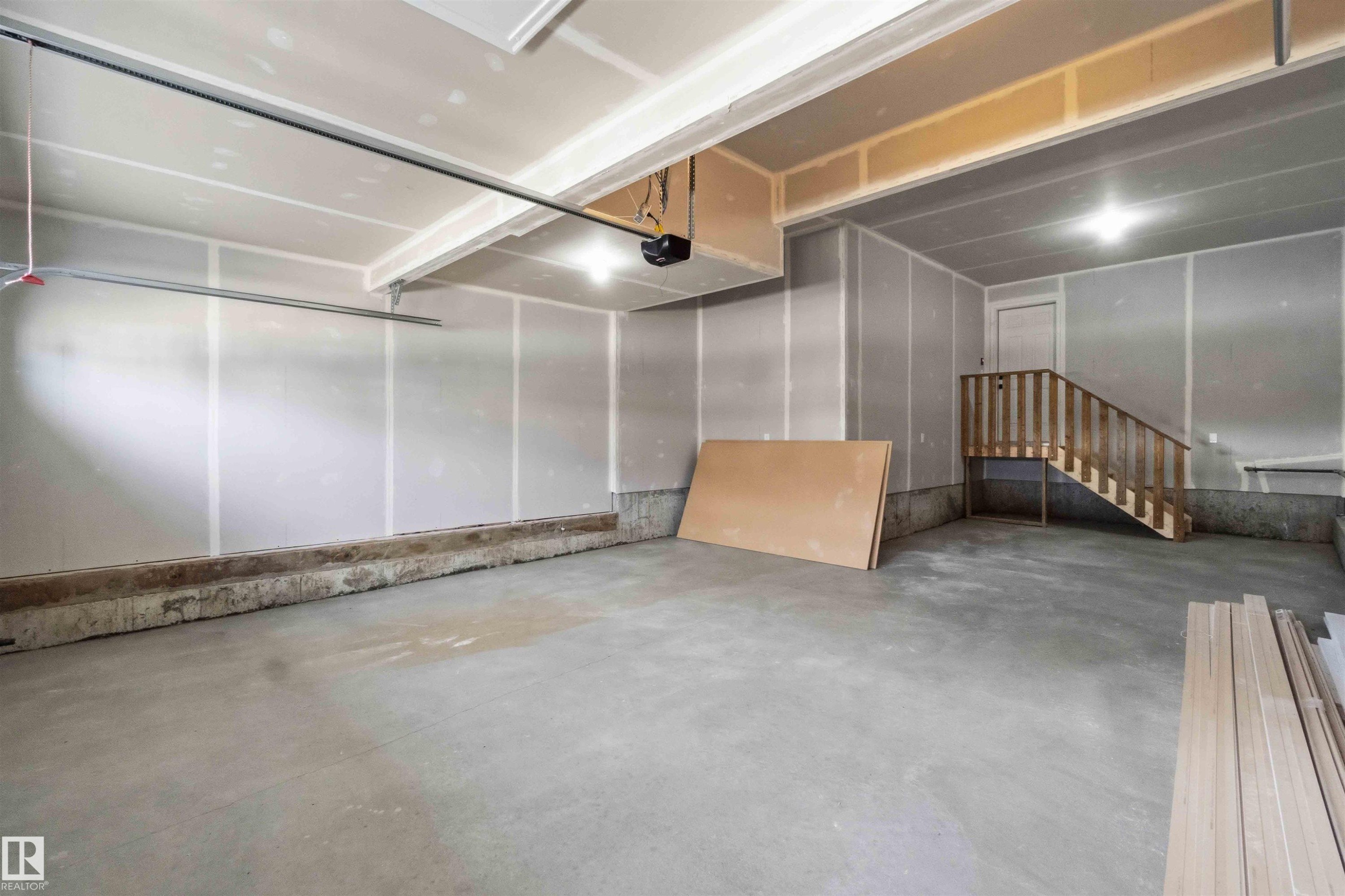 Spacious garage interior featuring a concrete floor, unfinished drywall, exposed ceiling beams, a wooden staircase, and built-in overhead lighting - 1085 Cristall Crescent Sw, Edmonton, AB - Indoor