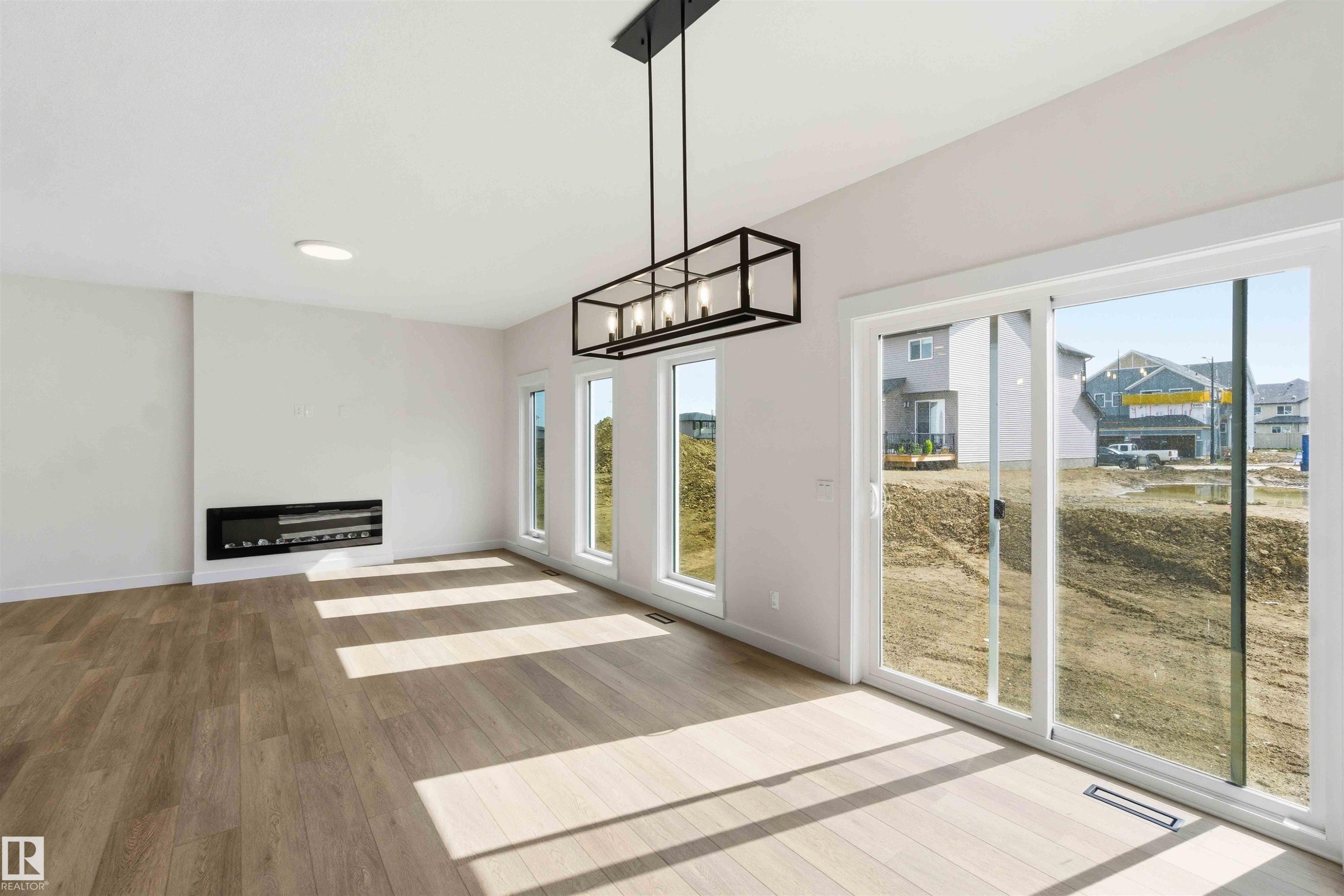 Spacious interior with wood-finish flooring, an electric fireplace, a linear chandelier, multiple windows, and a sliding glass door - 1085 Cristall Crescent Sw, Edmonton, AB - Indoor Photo Showing Other Room With Fireplace