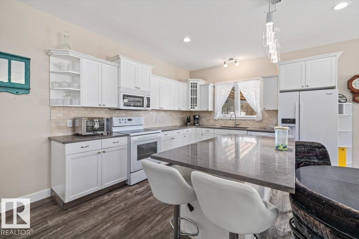 Modern kitchen featuring a central island with a stone-finish countertop, white cabinetry, integrated appliances, wood-finish flooring, and a tile backsplash - 4708 47A Avenue, Onoway, AB