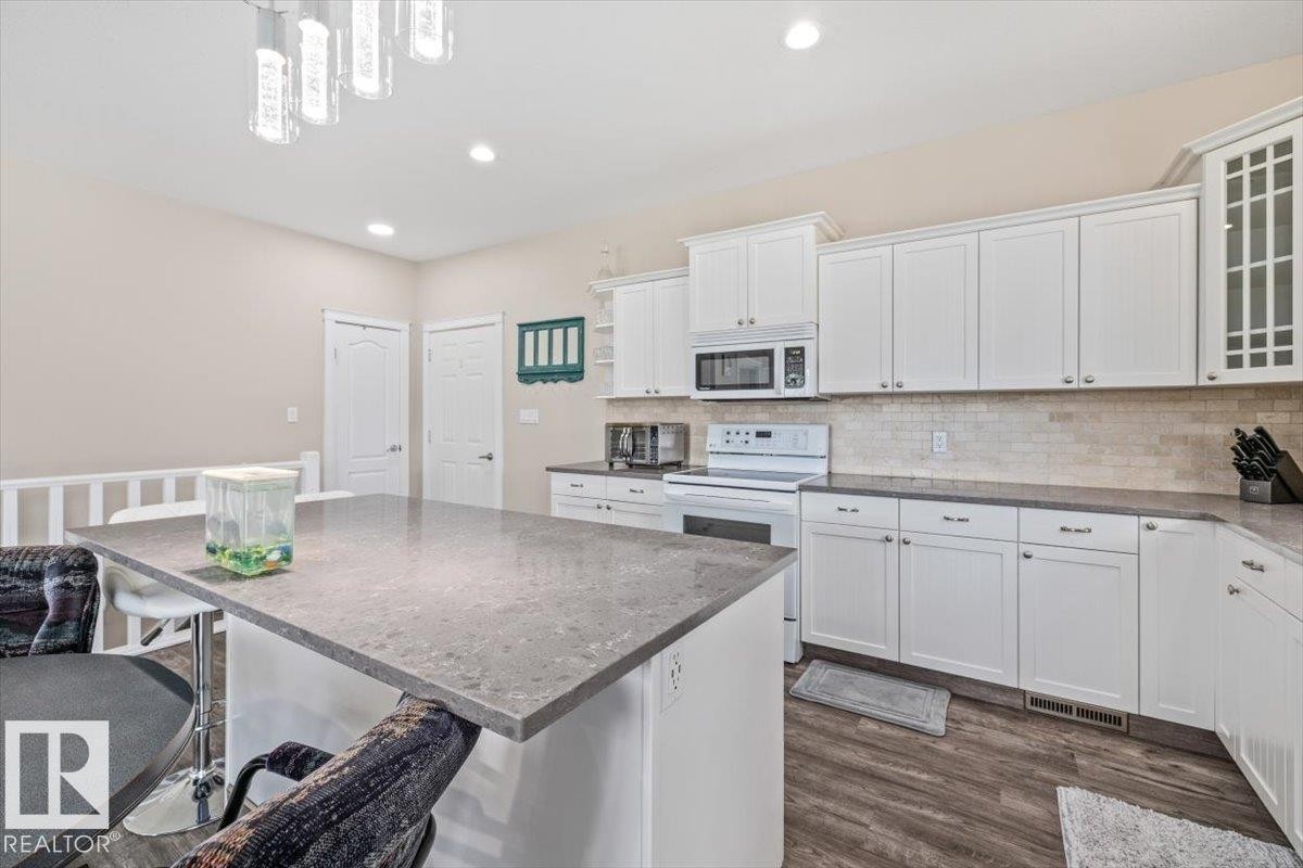 Modern kitchen featuring a large center island with a stone-finish countertop, white cabinetry, a tiled backsplash, wood-finish flooring, and recessed lighting - 4708 47A Avenue, Onoway, AB