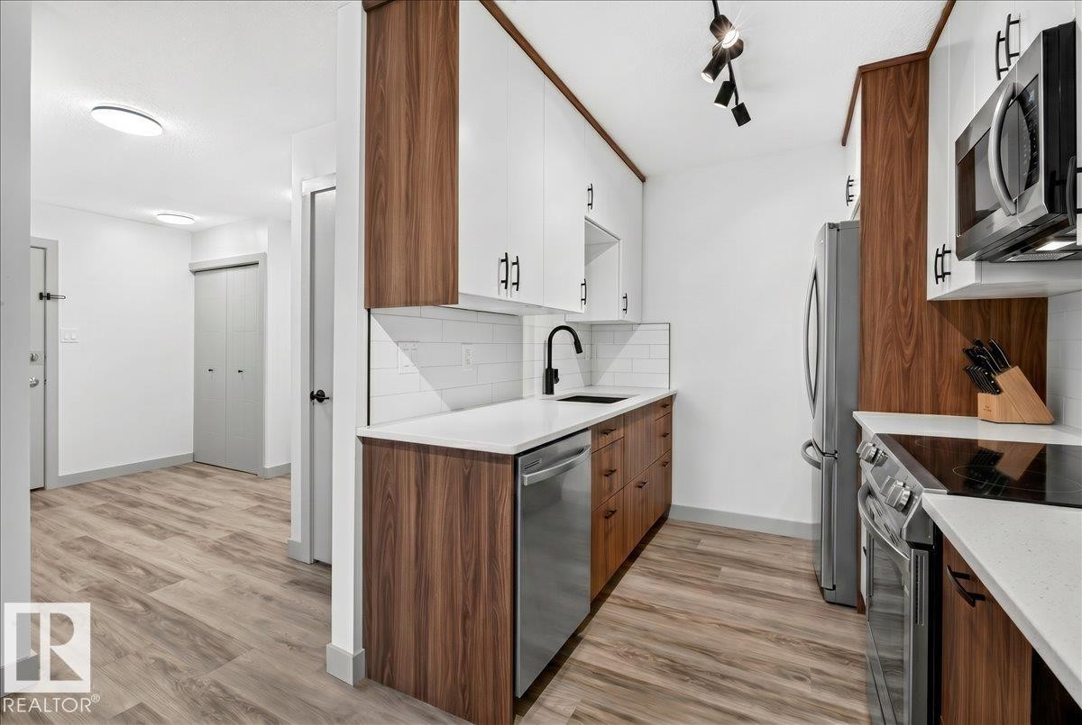 Modern kitchen featuring stainless steel appliances, wood-finish lower cabinetry, white upper cabinetry with matte black hardware, and a subway tile backsplash - 203 10624 123 Street, Edmonton, AB