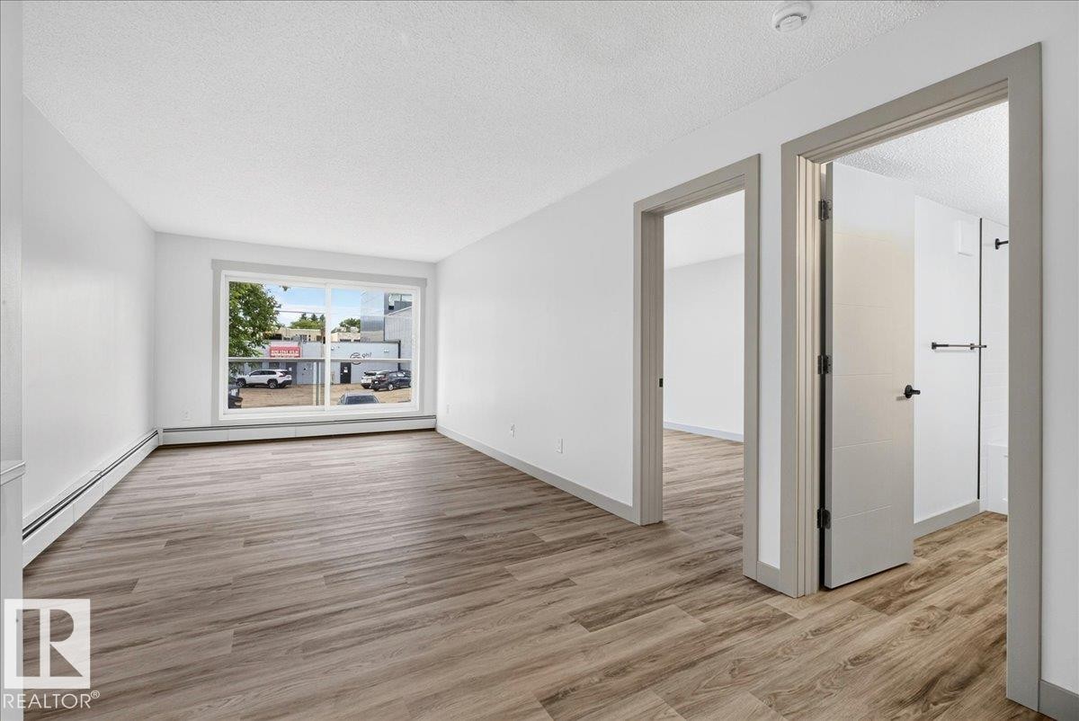 Spacious interior showcasing wood-finish flooring, white walls, and a large window - 203 10624 123 Street, Edmonton, AB