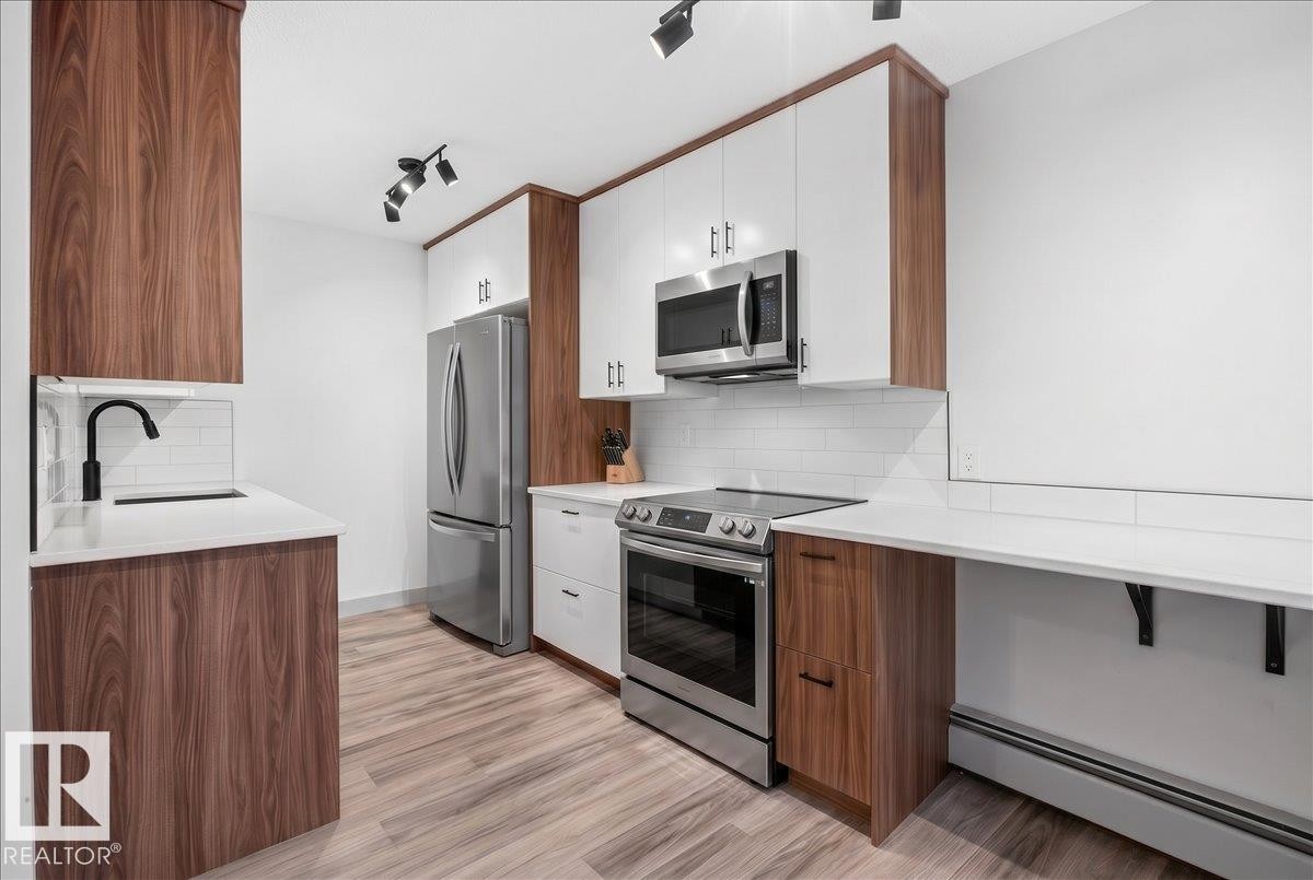 Modern kitchen featuring two-tone cabinetry, stainless steel appliances, a white subway tile backsplash, white countertops, and wood-finish flooring - 203 10624 123 Street, Edmonton, AB