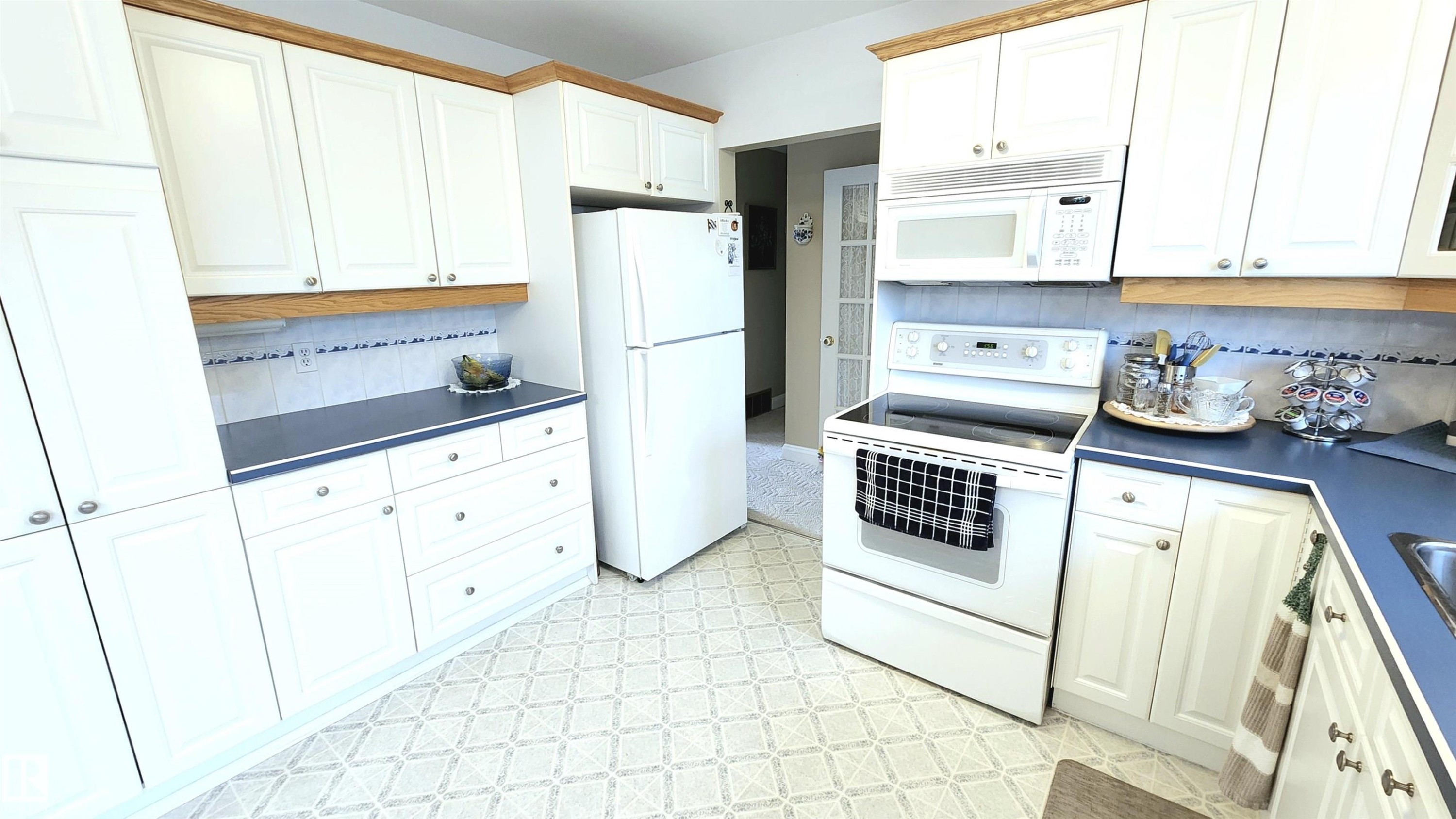 Kitchen featuring white cabinetry with brushed nickel hardware, laminate countertops, white tile backsplash with decorative accent, and patterned resilient flooring - 13307 Delwood Road, Edmonton, AB