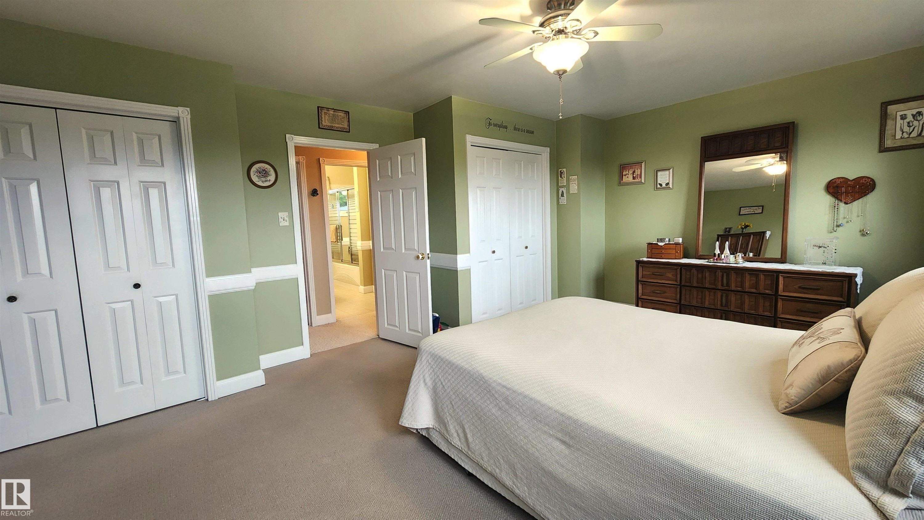 Spacious bedroom featuring neutral carpeting, two sets of bi-fold closet doors, chair rail molding, and an overhead ceiling fan with integrated lighting - 13307 Delwood Road, Edmonton, AB