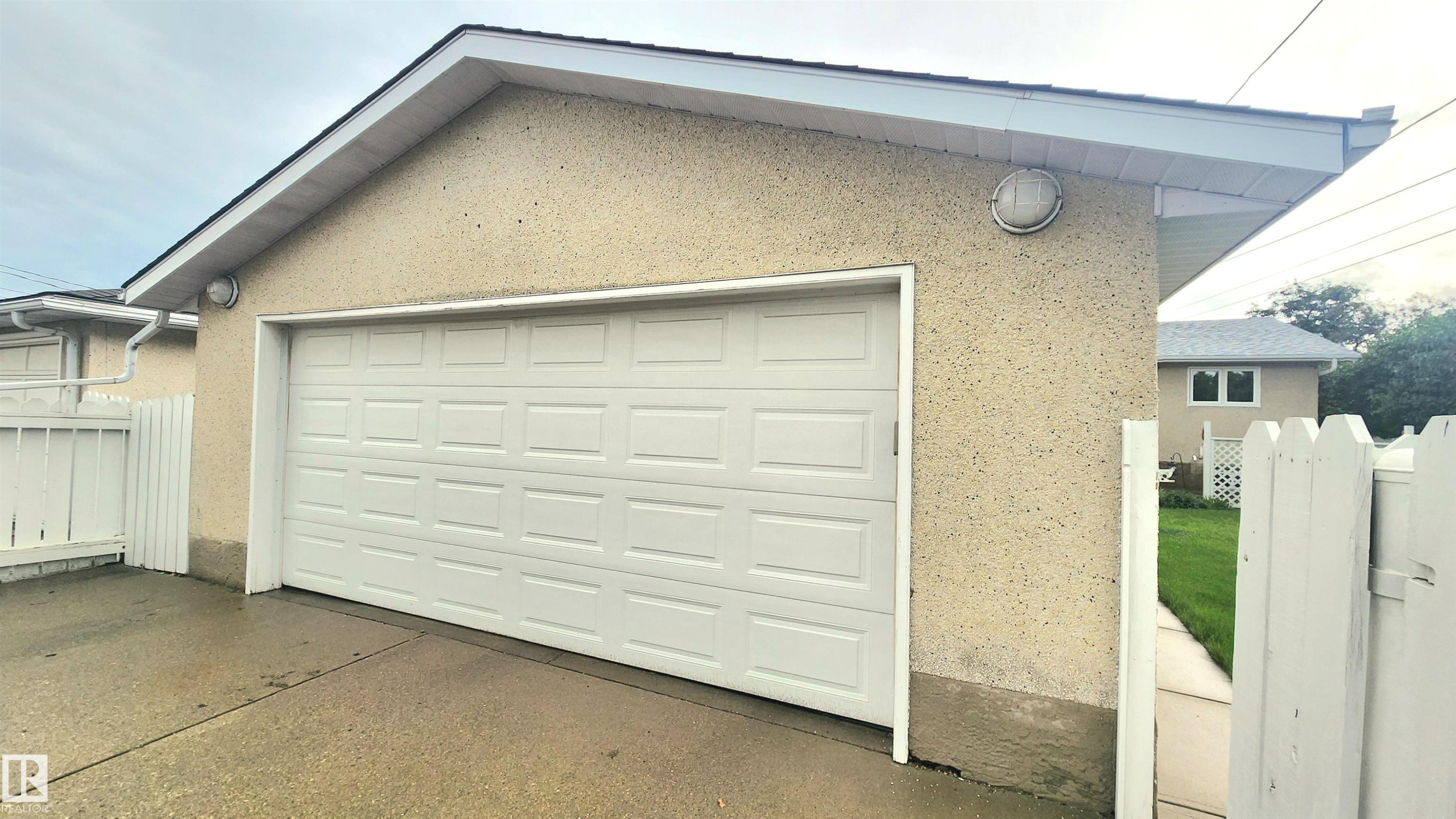 Detached garage featuring a paneled overhead door, stucco exterior, and white picket fence - 13307 Delwood Road, Edmonton, AB
