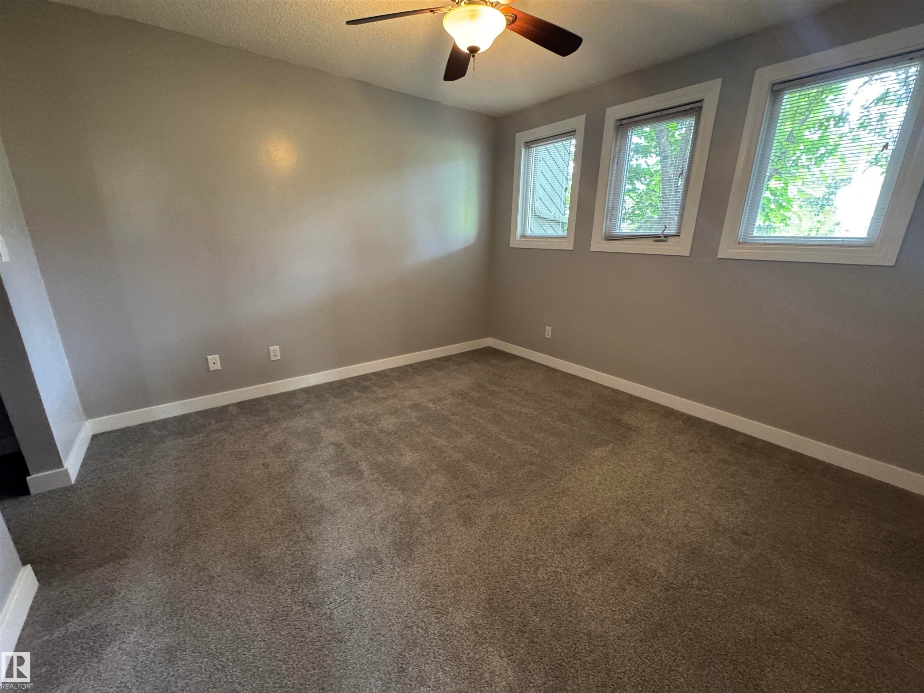 Carpeted room with a ceiling fan light fixture, three windows, and neutral wall paint - 6441 180 Street, Edmonton, AB