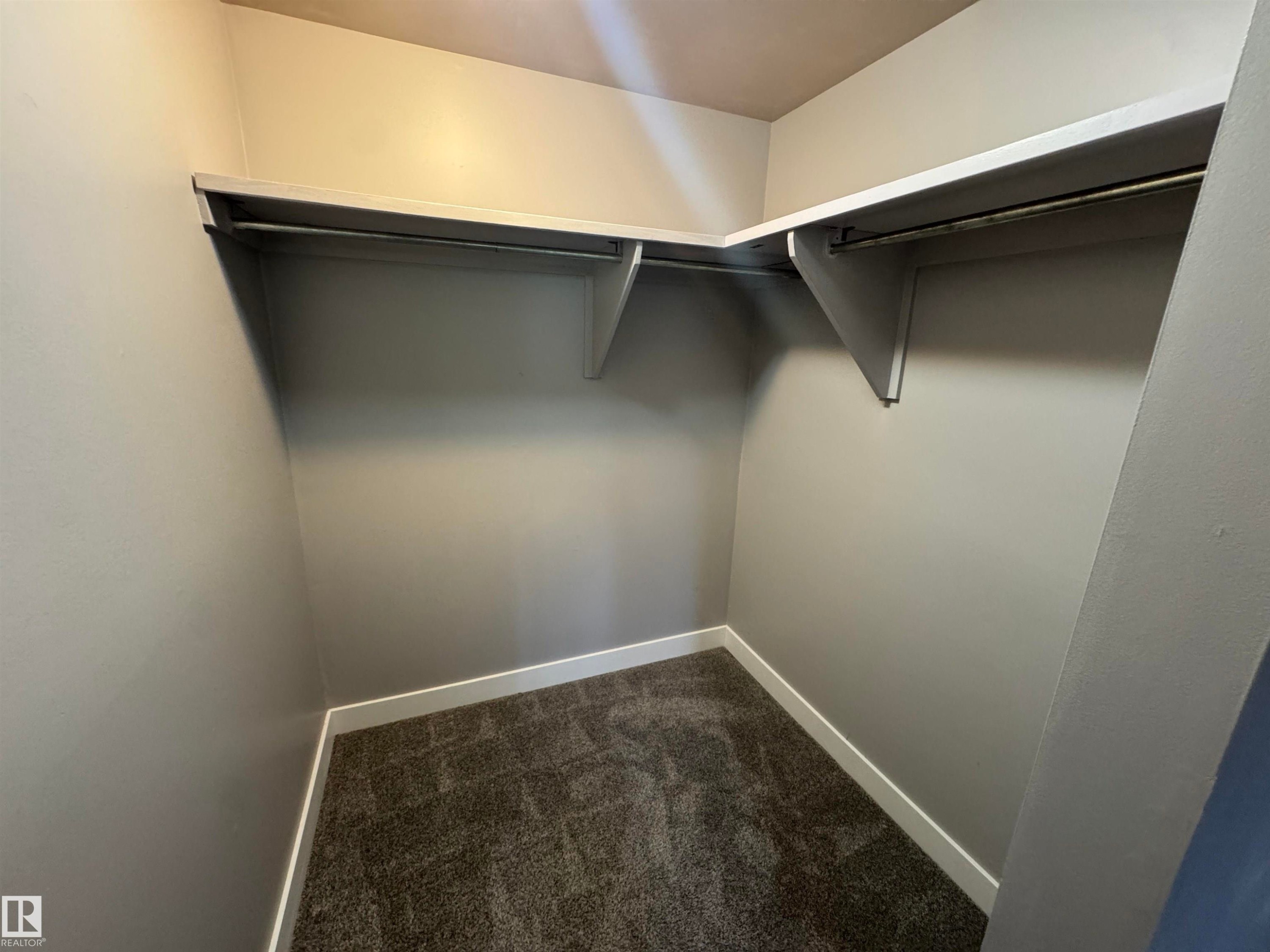 Walk-in closet featuring L-shaped shelving, clothing rods, light grey wall paint, white baseboards, and dark grey patterned carpeting - 6441 180 Street, Edmonton, AB