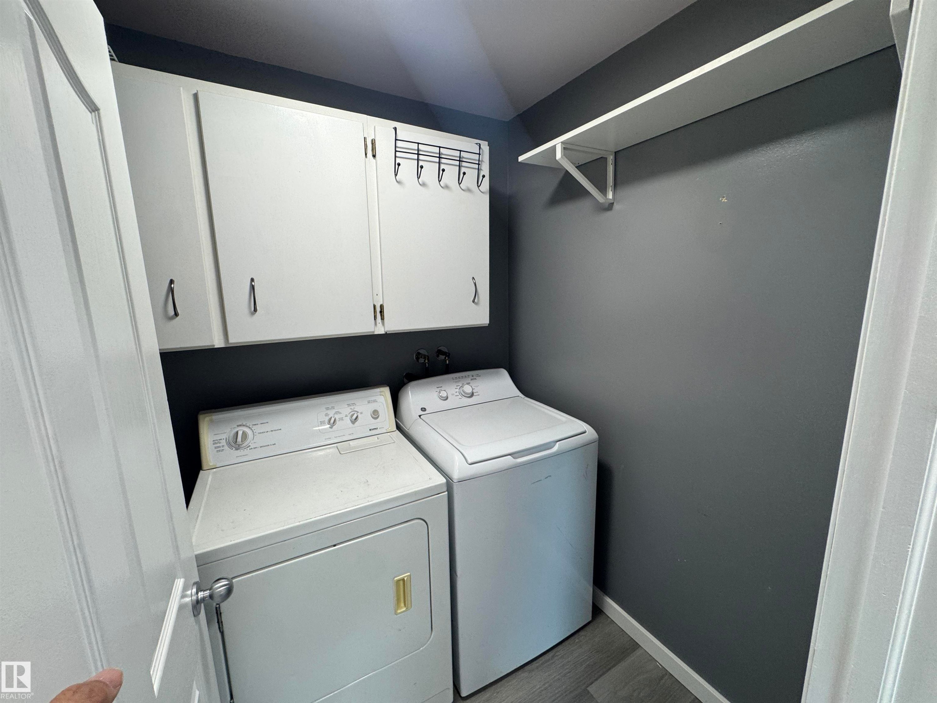 Dedicated laundry area featuring overhead white cabinetry, a wall-mounted shelving unit, and wood-finish flooring - 6441 180 Street, Edmonton, AB
