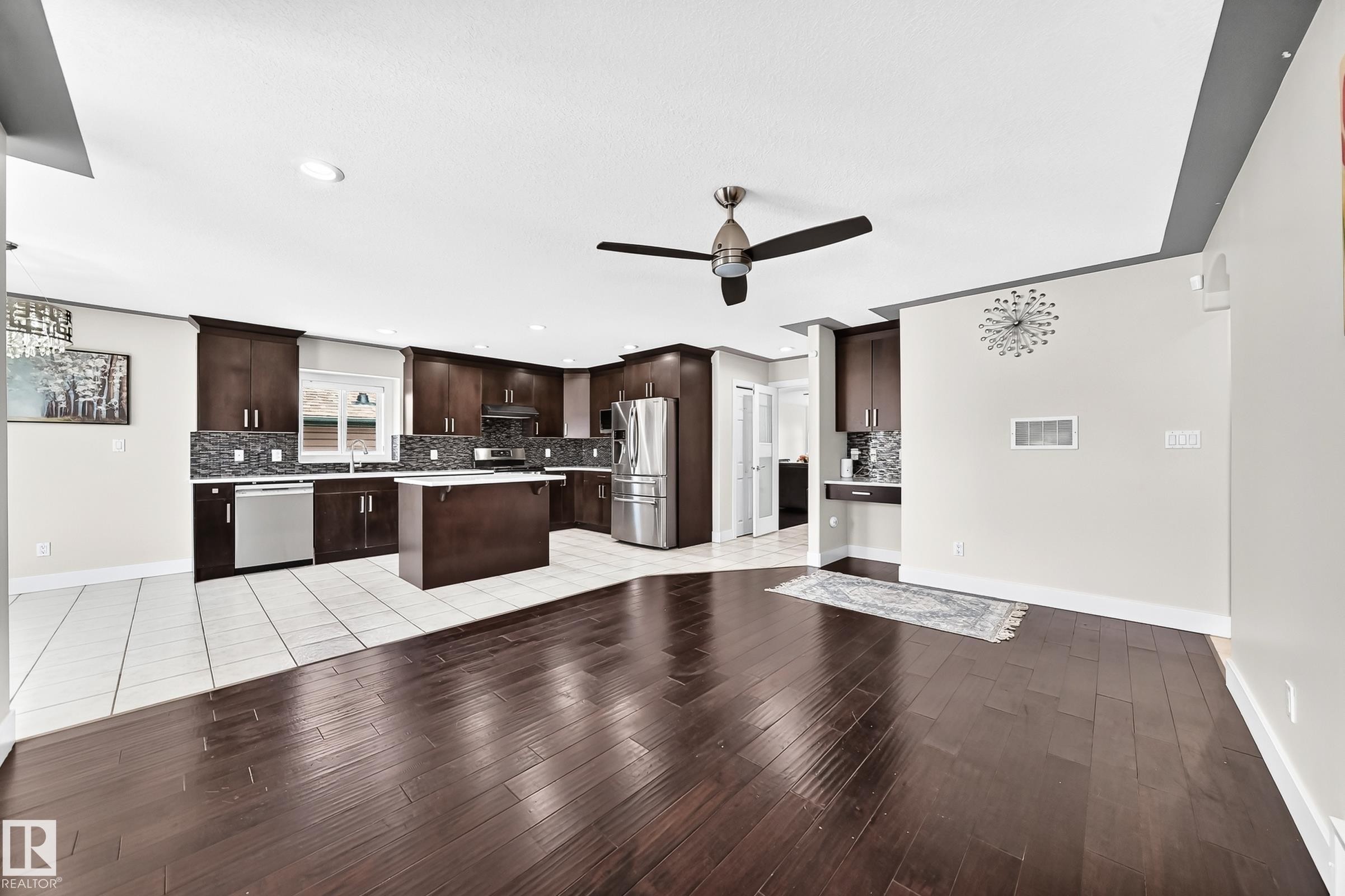 Open-concept kitchen and living area featuring rich wood-finish flooring, a contemporary ceiling fan, dark wood cabinetry, a mosaic tile backsplash, and stainless steel appliances - 16124 76 Street, Edmonton, AB