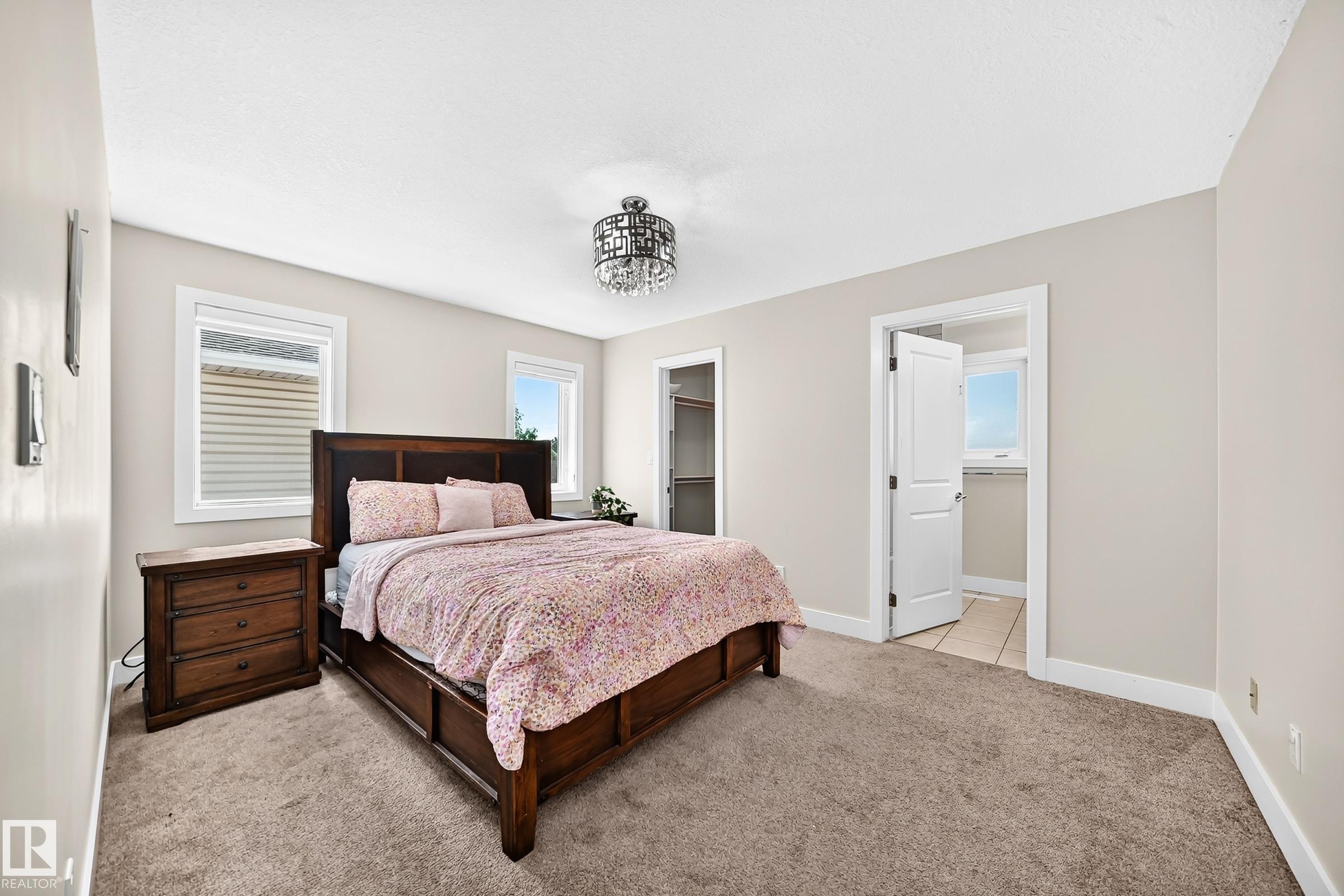 Carpeted room featuring two windows with horizontal blinds, a ceiling-mounted chandelier, and light-toned walls - 16124 76 Street, Edmonton, AB