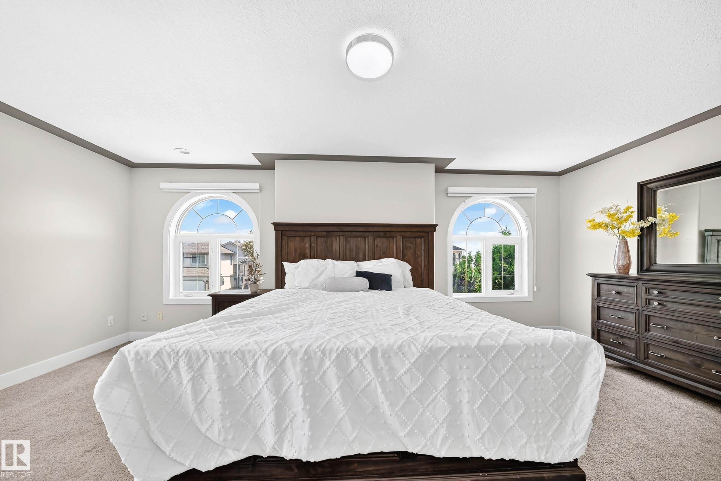 Carpeted bedroom featuring two arched windows, a dark wood-finish headboard, tray ceiling detail, and crown molding - 16124 76 Street, Edmonton, AB