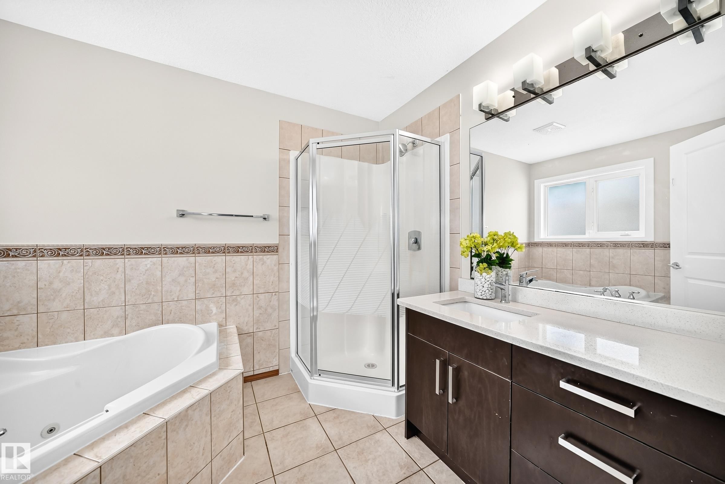 Full bathroom featuring a corner shower enclosure, a jetted soaking tub with tiled surround, a vanity with a white countertop and dark wood-finish cabinetry, and a large mirror with a multi-bulb light fixture - 16124 76 Street, Edmonton, AB