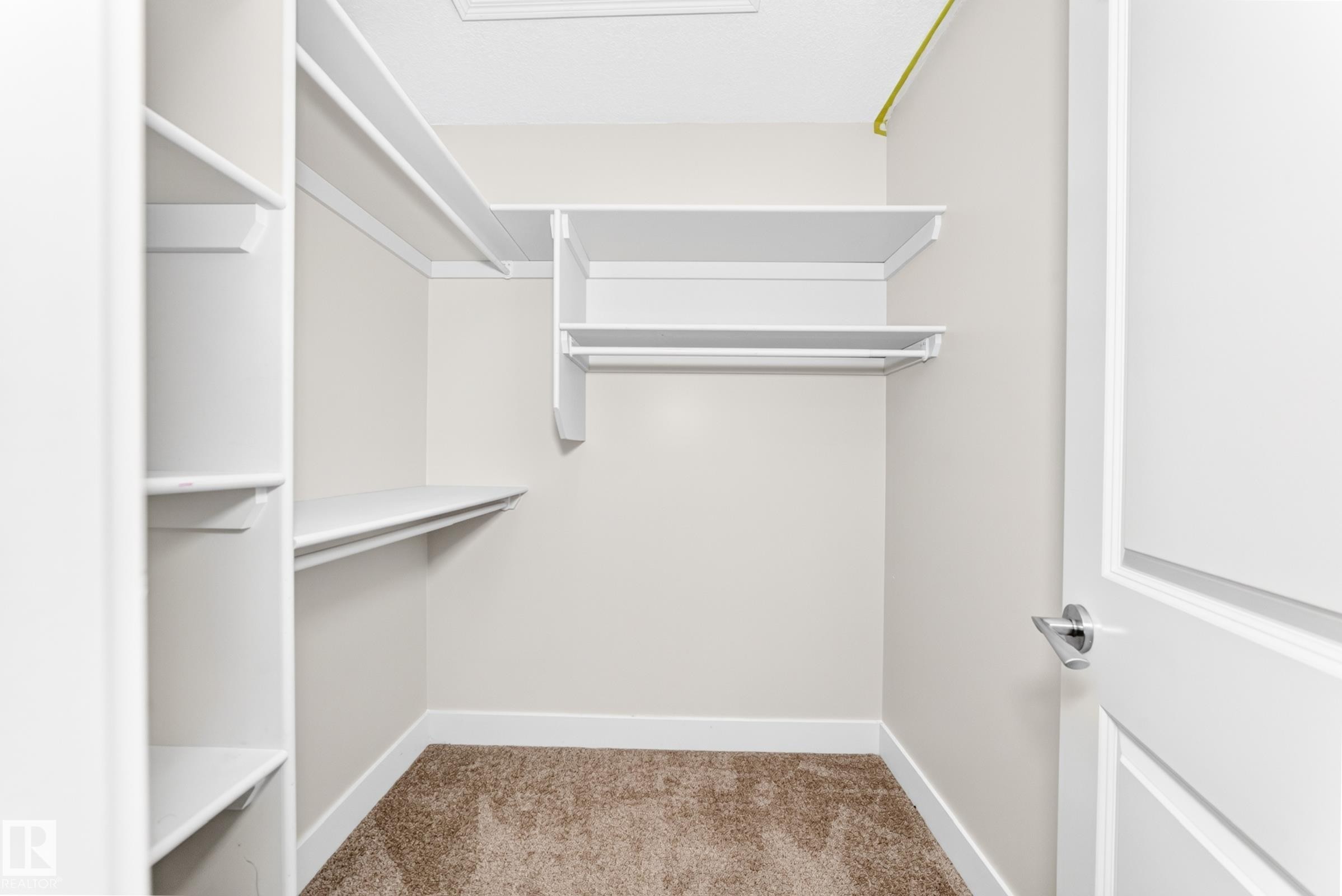 Walk-in closet featuring white shelving, multiple hanging rods, warm neutral wall paint, white trim, and brown carpet flooring - 16124 76 Street, Edmonton, AB