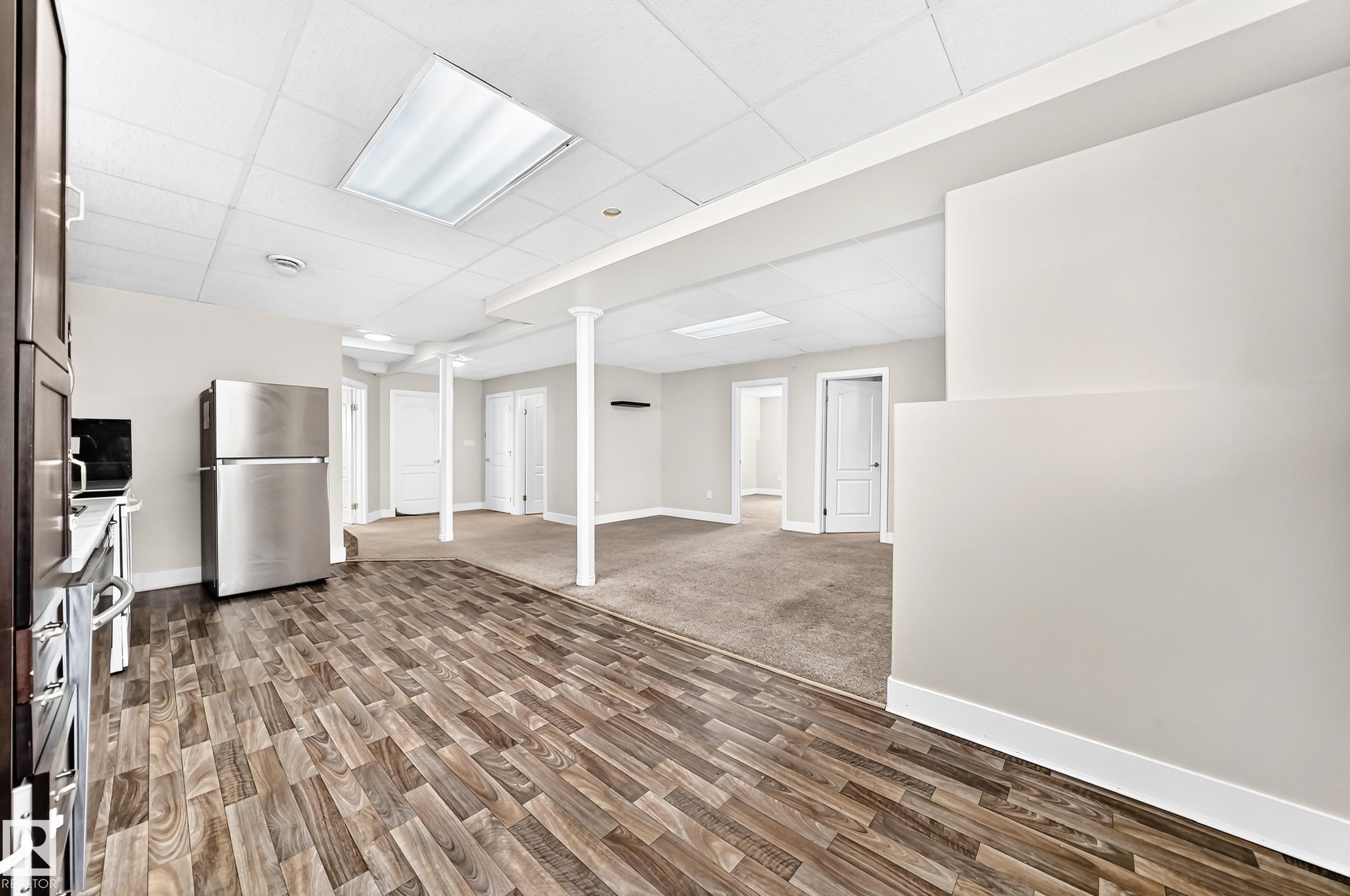 Lower level featuring wood-finish flooring, a stainless steel refrigerator, white paneled doors, exposed support columns, and recessed ceiling lighting - 16124 76 Street, Edmonton, AB