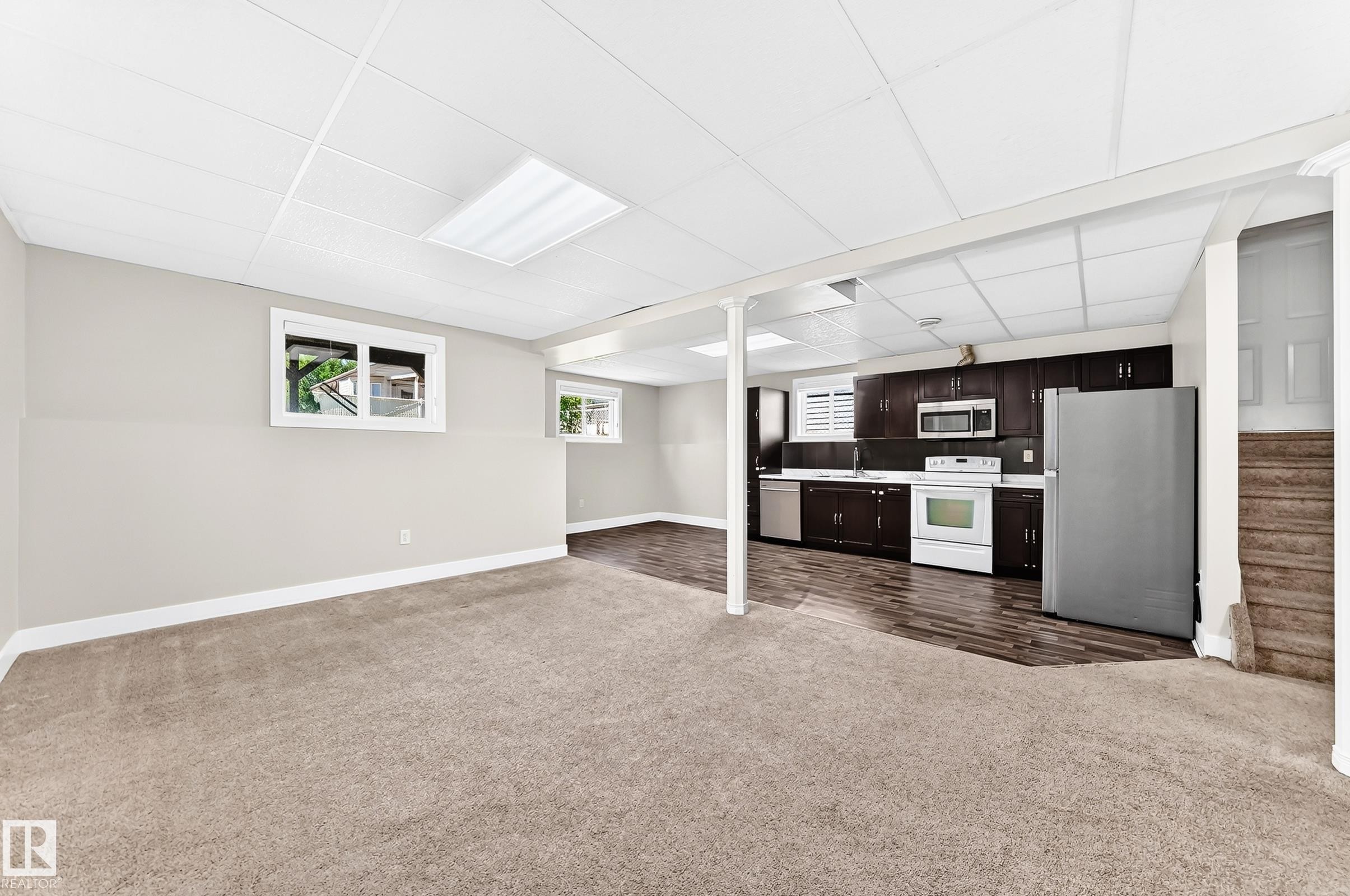 Lower level living area featuring a full kitchen with dark wood-finish cabinetry, stainless steel refrigerator, white range, and built-in microwave - 16124 76 Street, Edmonton, AB