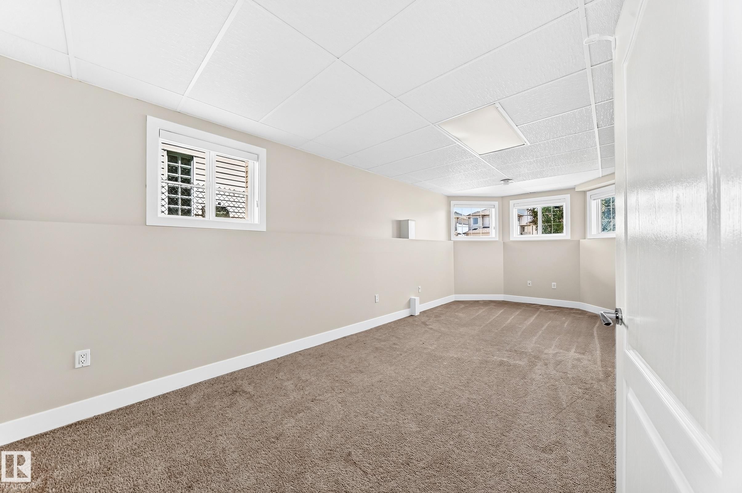 Spacious carpeted room featuring multiple windows, a drop ceiling with recessed lighting, light-toned walls, and white trim - 16124 76 Street, Edmonton, AB