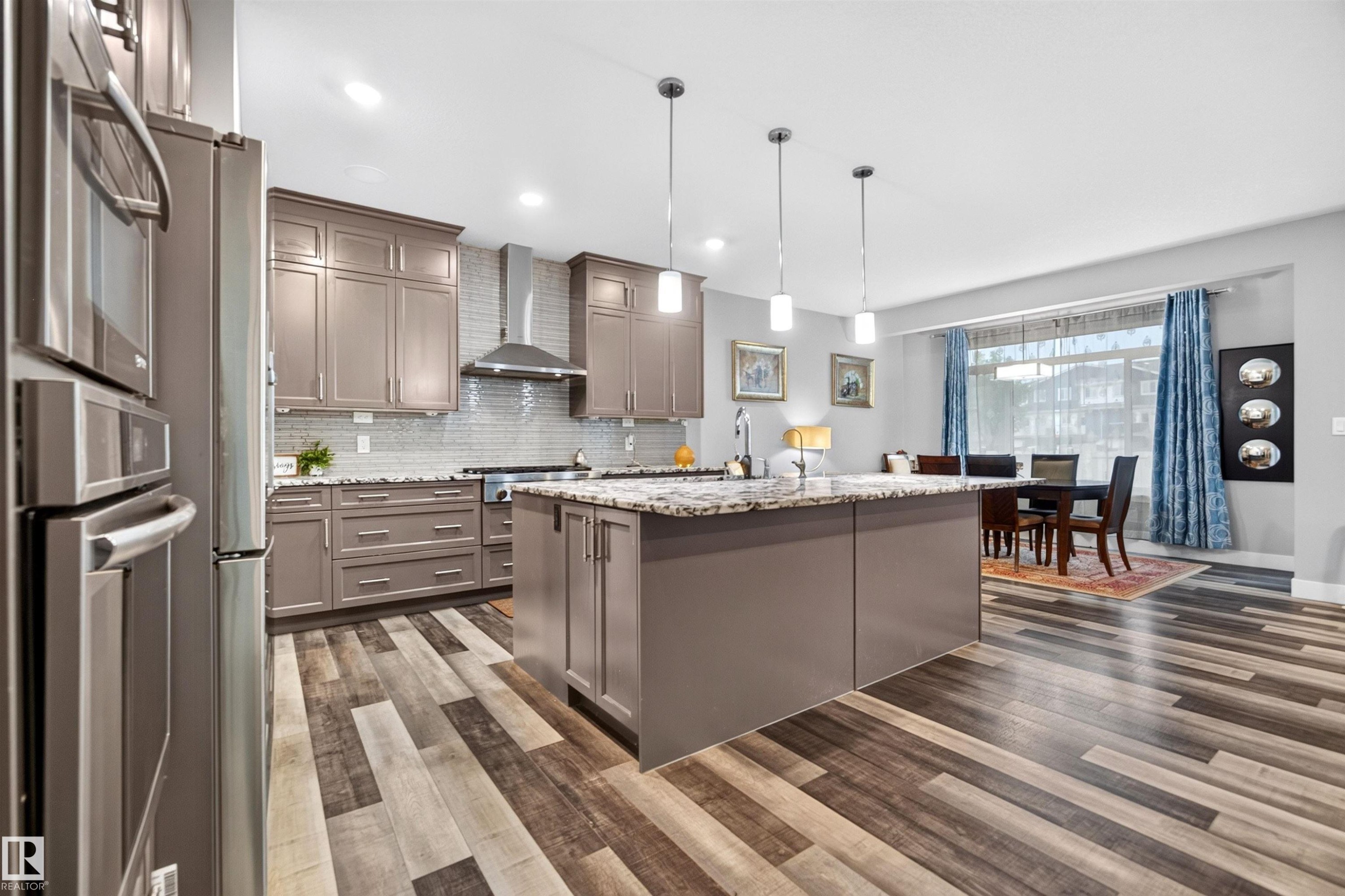Open-concept kitchen featuring wood-finish flooring, a large island with pendant lighting, stainless steel appliances, taupe cabinetry, and granite countertops - 1603 158 Street, Edmonton, AB