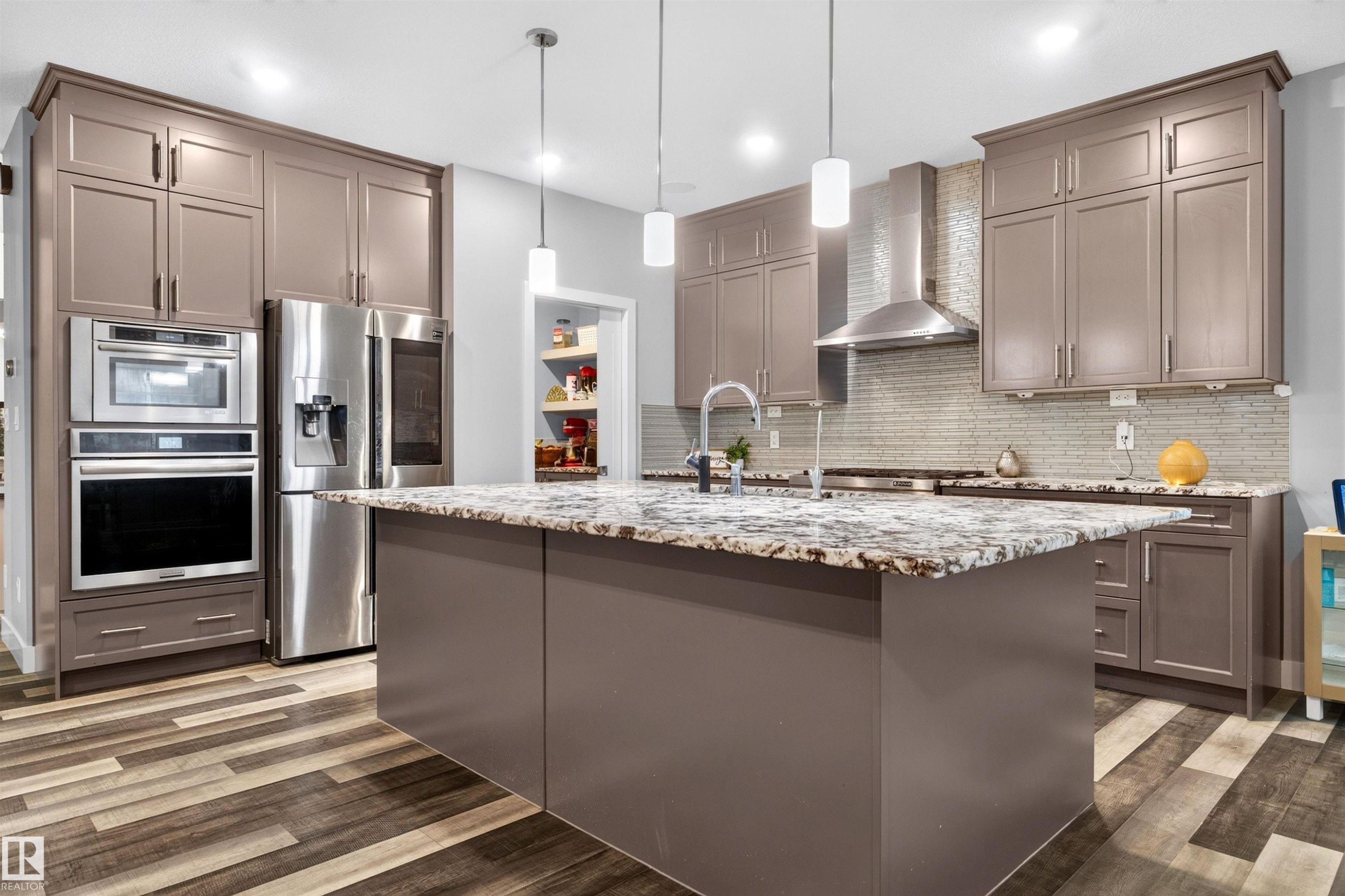 Spacious kitchen featuring a large island with a stone countertop, extensive cabinetry, stainless steel appliances, a tile backsplash, and wood-finish flooring - 1603 158 Street, Edmonton, AB