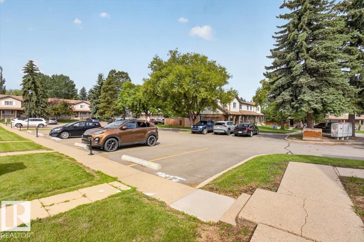 Paved parking area surrounded by mature trees and grass, with multi-story residential buildings in the background - 27 Walden Park, St. Albert, AB