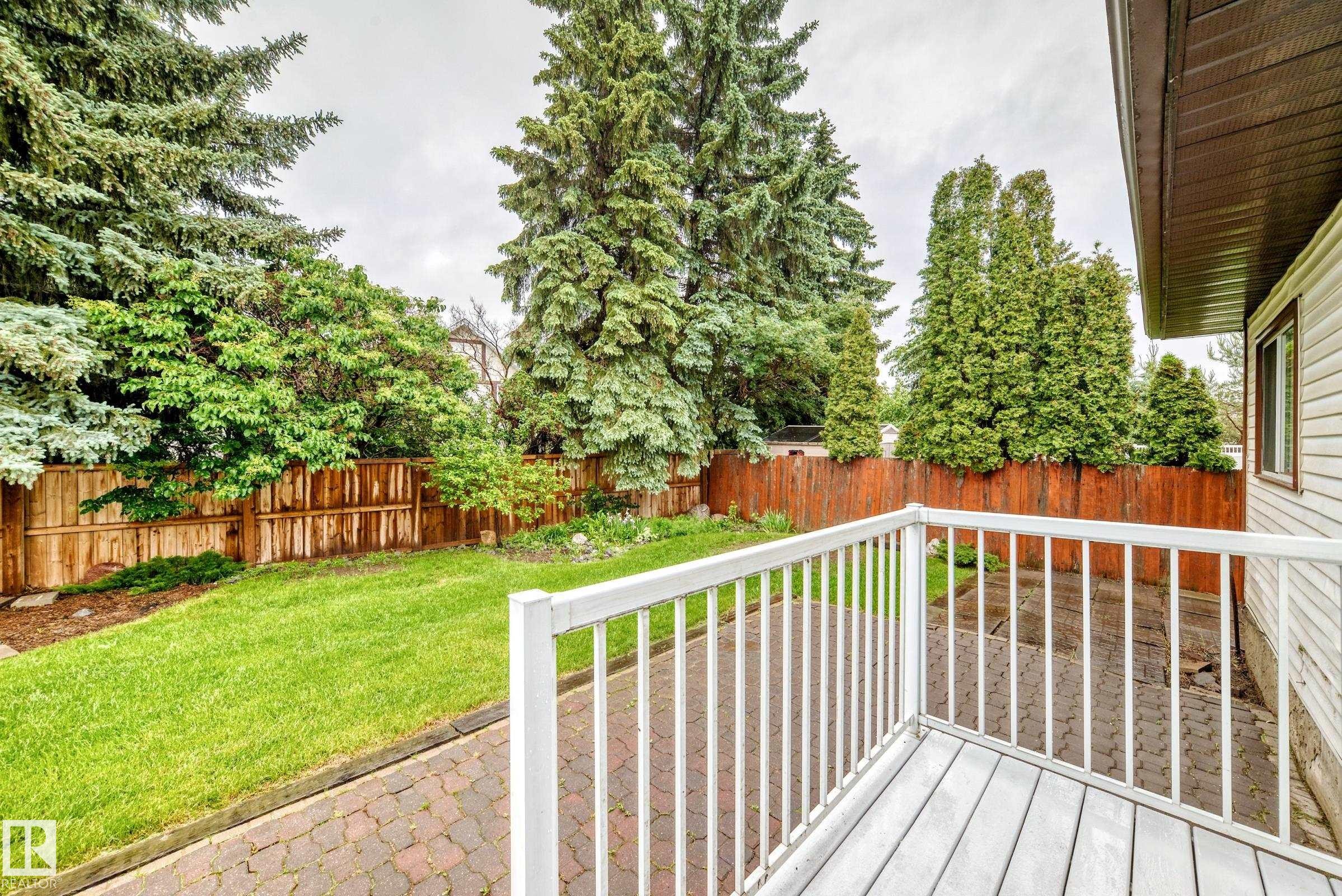 Outdoor deck with white railing, wood-finish flooring, a paved patio, a manicured lawn, and mature perimeter trees - 9843 188 Street, Edmonton, AB