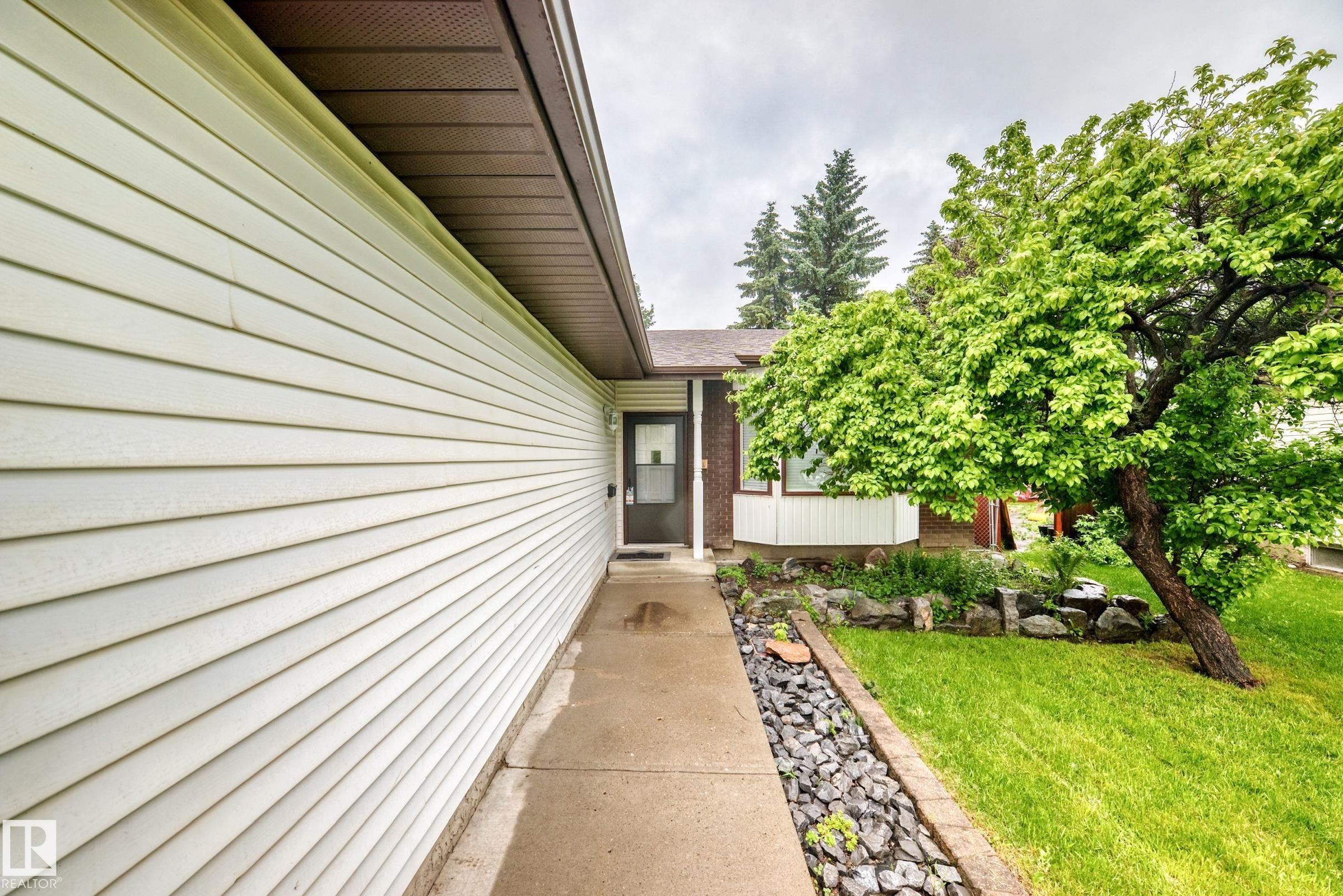 Exterior featuring light-colored siding, a concrete walkway, established landscaping with a mature tree, and a dark entry door - 9843 188 Street, Edmonton, AB