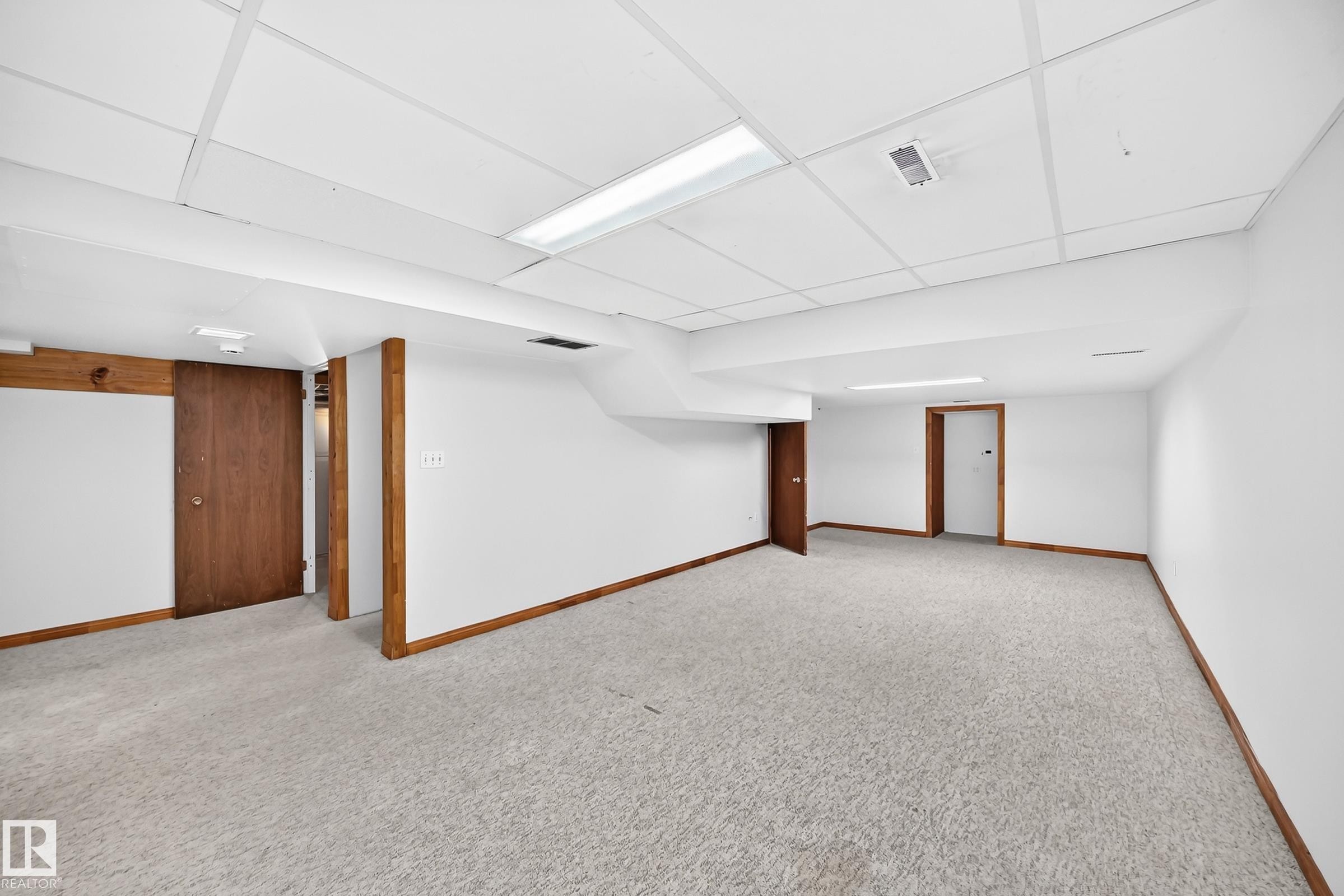 Spacious lower-level room featuring light-toned carpeting, white walls, wood-finish trim, and a drop ceiling with integrated fluorescent lighting - 9843 188 Street, Edmonton, AB