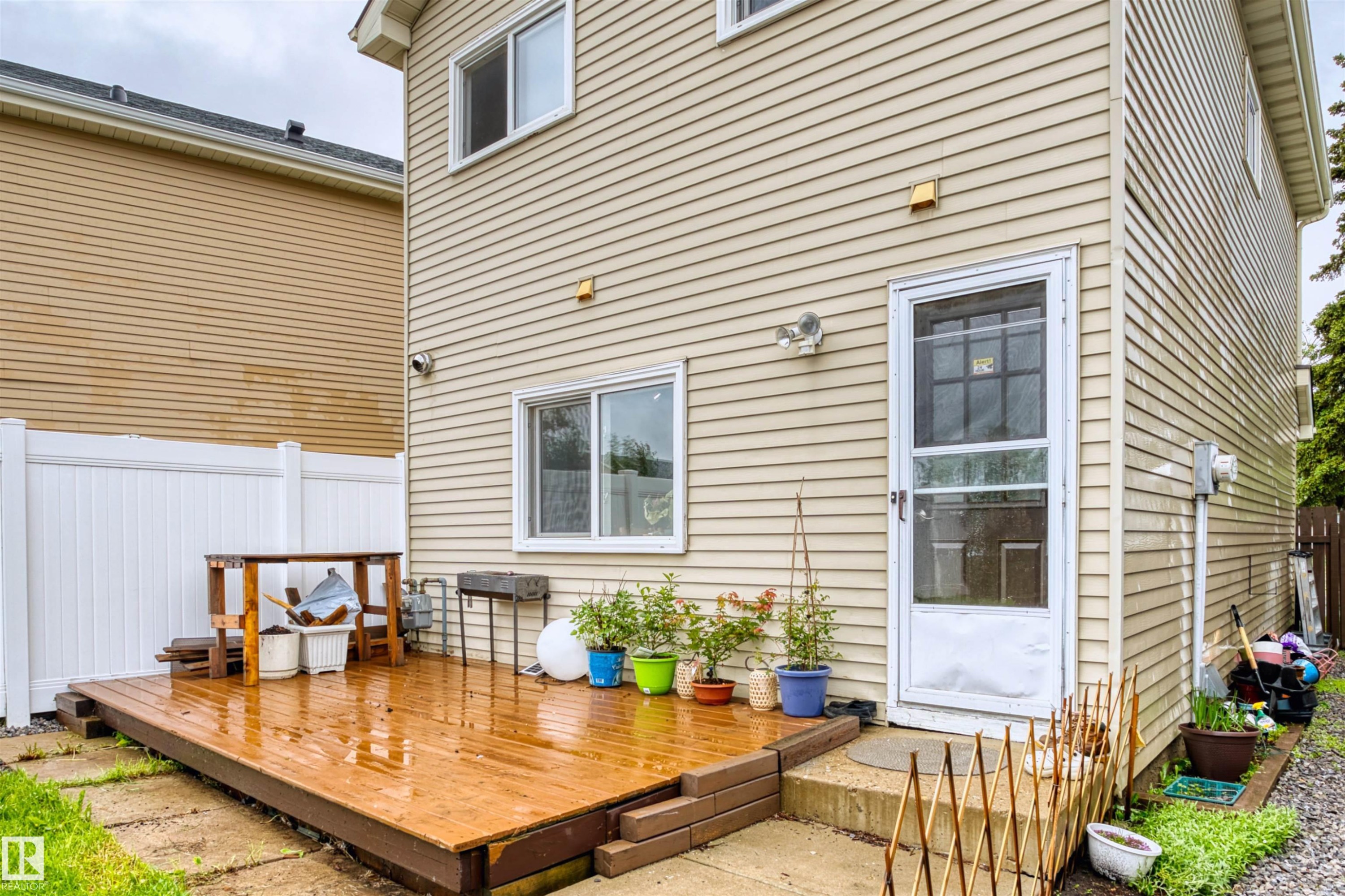 Rear exterior featuring beige horizontal siding, a ground-level wood-finish deck, white vinyl fencing, a white exterior door with a glass storm door, and a white-framed window - 359 Kirkpatrick Crescent, Edmonton, AB