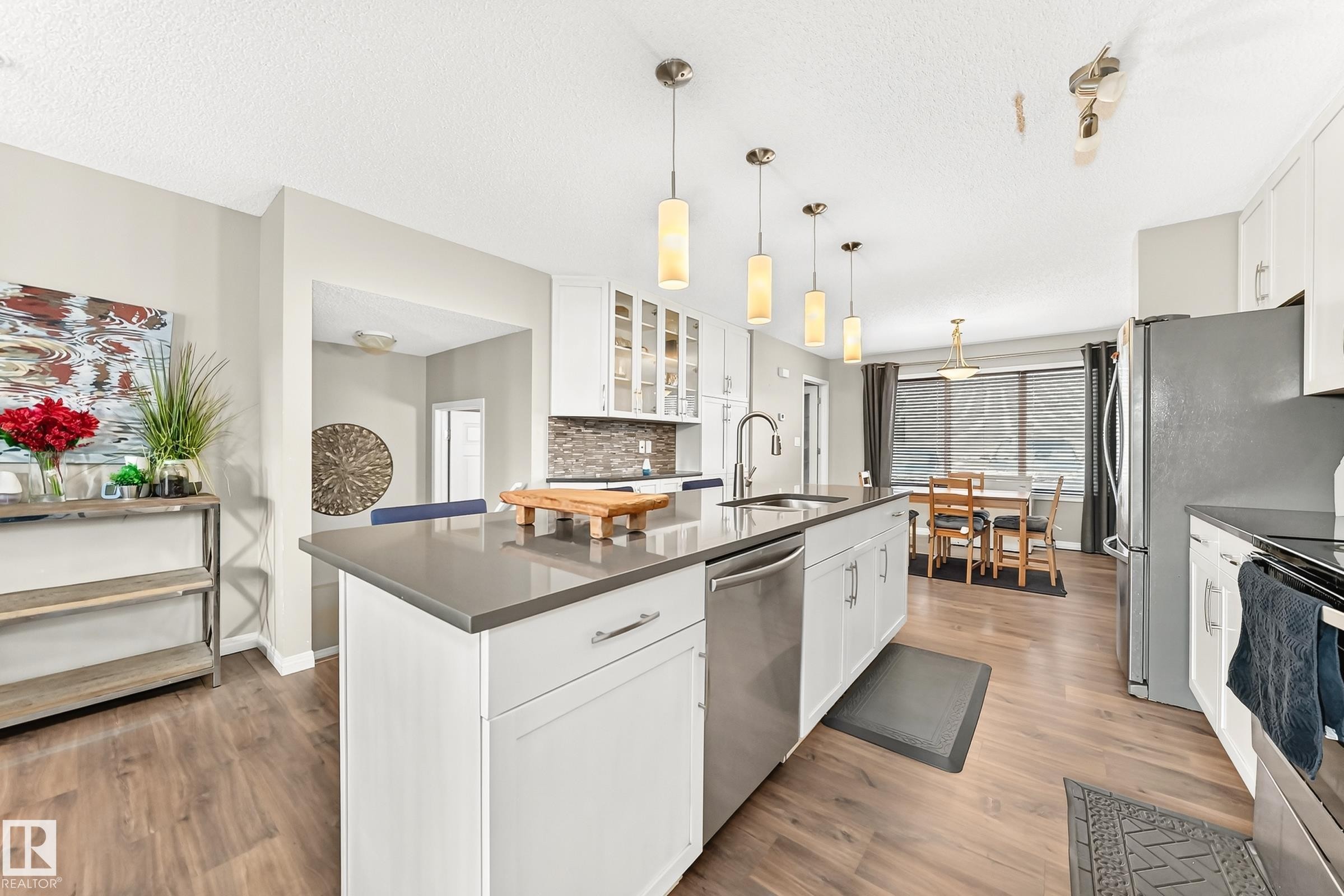 Kitchen featuring an island with a built-in sink, stainless steel appliances, white cabinetry, wood-finish flooring, and pendant lighting - 875 Chappelle Link Link Sw, Edmonton, AB