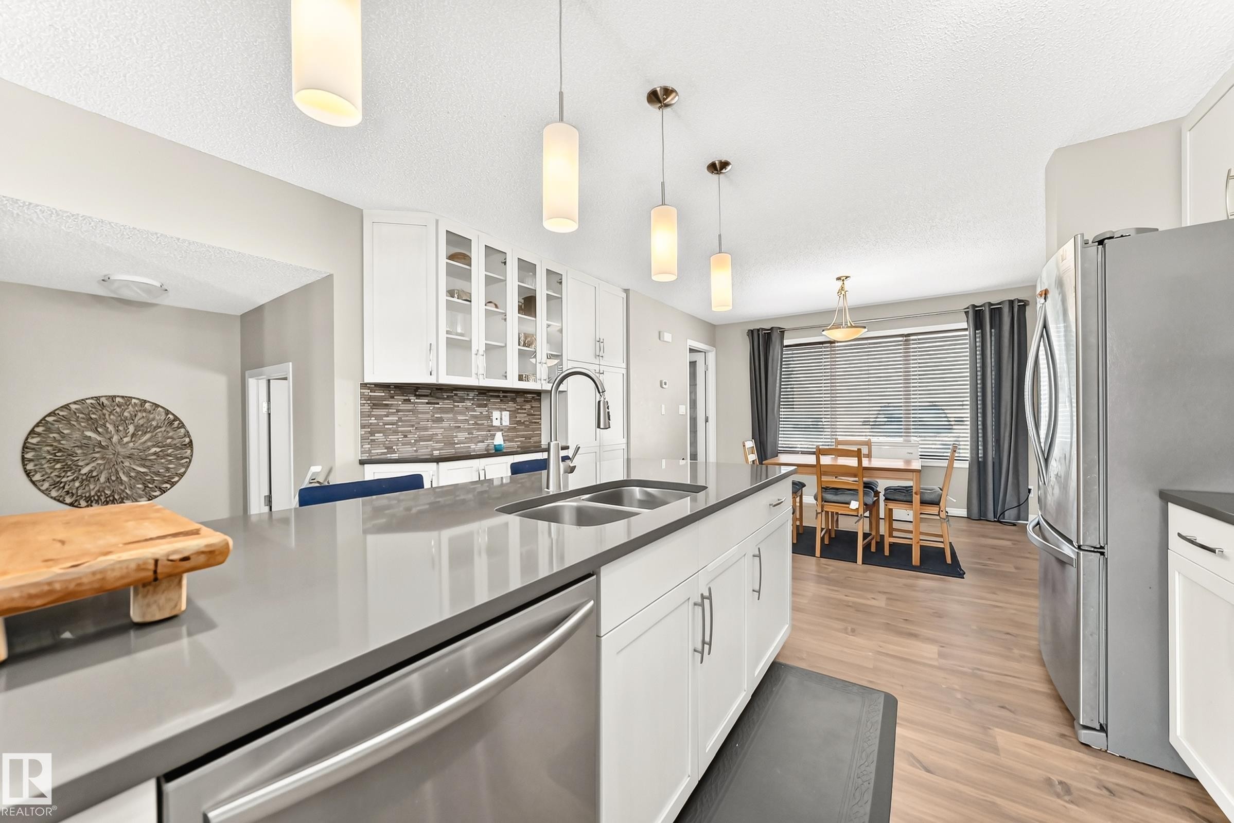 Kitchen featuring a large island with dual sinks, stainless steel appliances, white cabinetry, and wood-finish flooring - 875 Chappelle Link Link Sw, Edmonton, AB