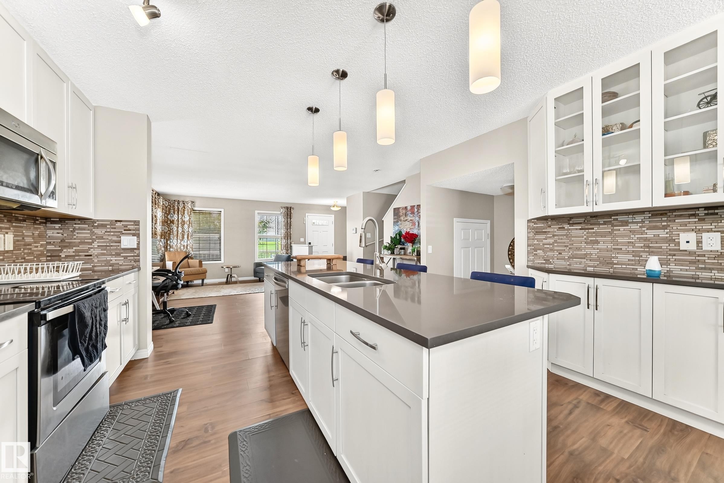 Kitchen featuring a large center island with integrated sink, stainless steel appliances, wood-finish flooring, white cabinetry, and a patterned tile backsplash - 875 Chappelle Link Link Sw, Edmonton, AB