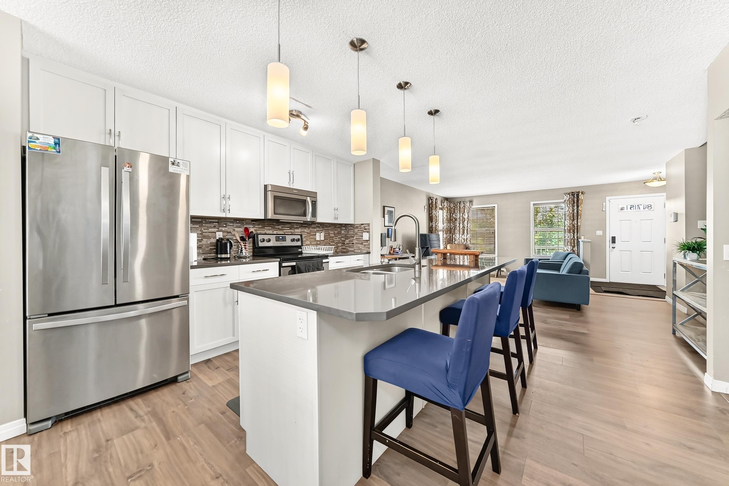 Kitchen featuring an expansive island with a countertop, pendant lighting, stainless steel appliances, white cabinetry, and wood-finish flooring - 875 Chappelle Link Link Sw, Edmonton, AB