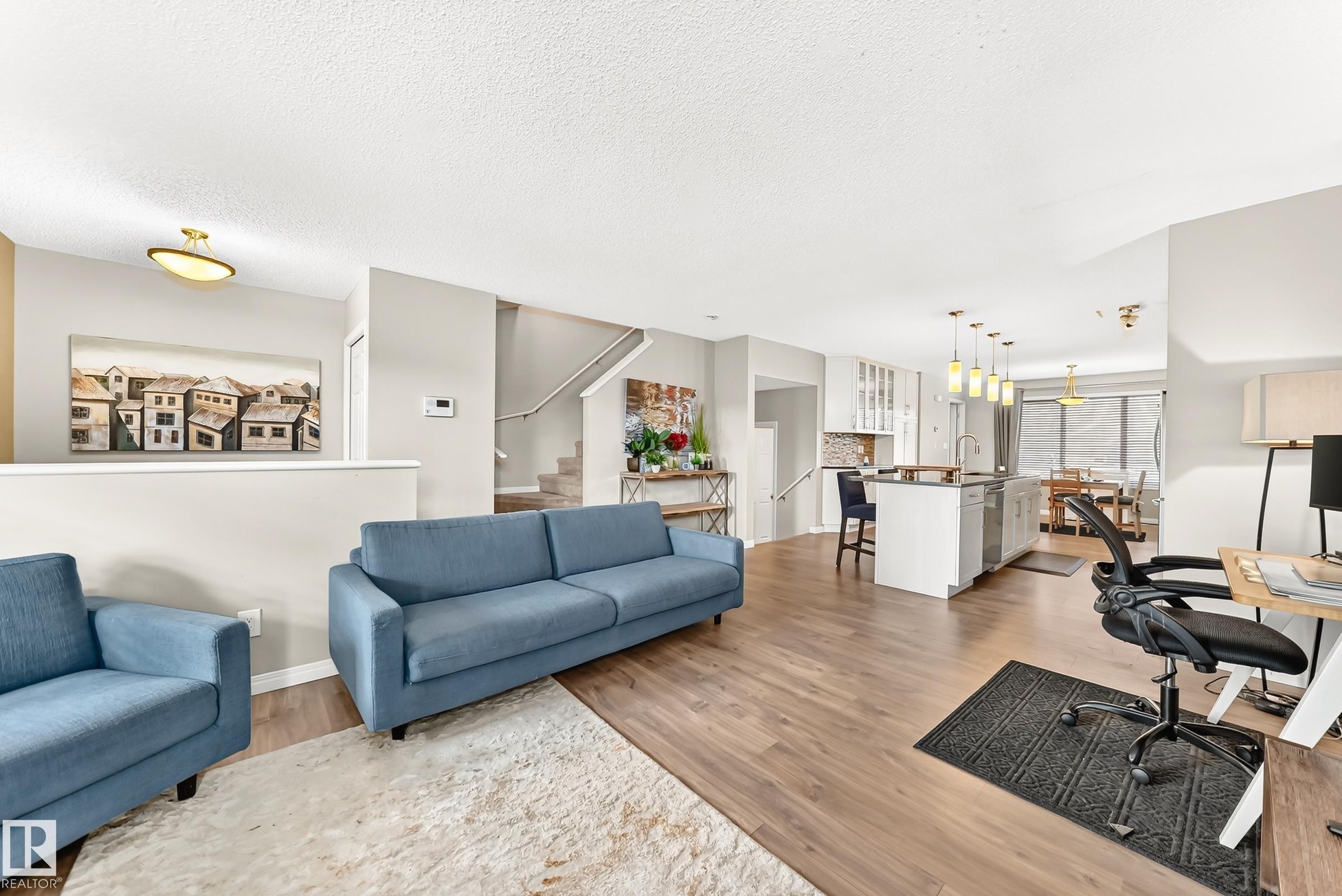 Spacious open-concept living area featuring wood-finish flooring, a contemporary kitchen island with pendant lighting, white cabinetry, and a full stainless steel appliance suite - 875 Chappelle Link Link Sw, Edmonton, AB