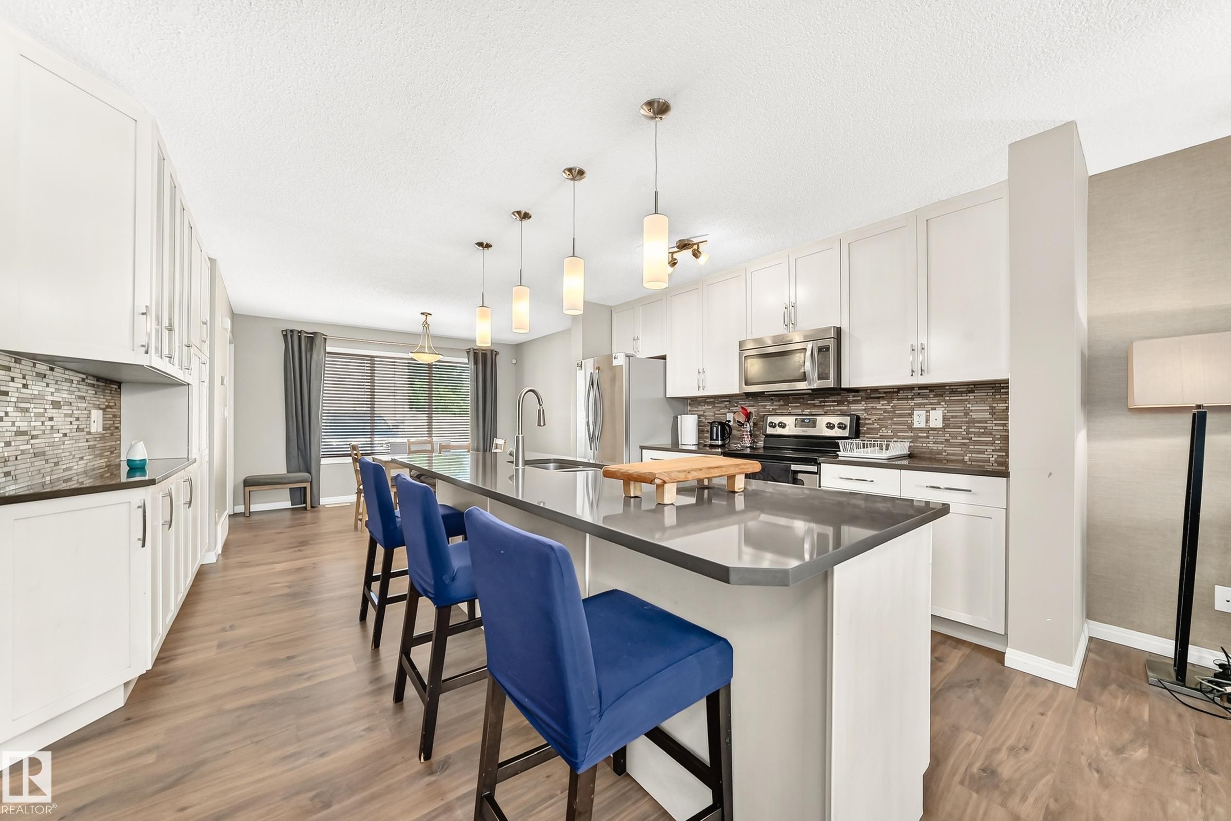 Open-concept kitchen featuring an elongated island with seating, stainless steel appliances, white cabinetry, wood-finish flooring, and pendant lighting - 875 Chappelle Link Link Sw, Edmonton, AB
