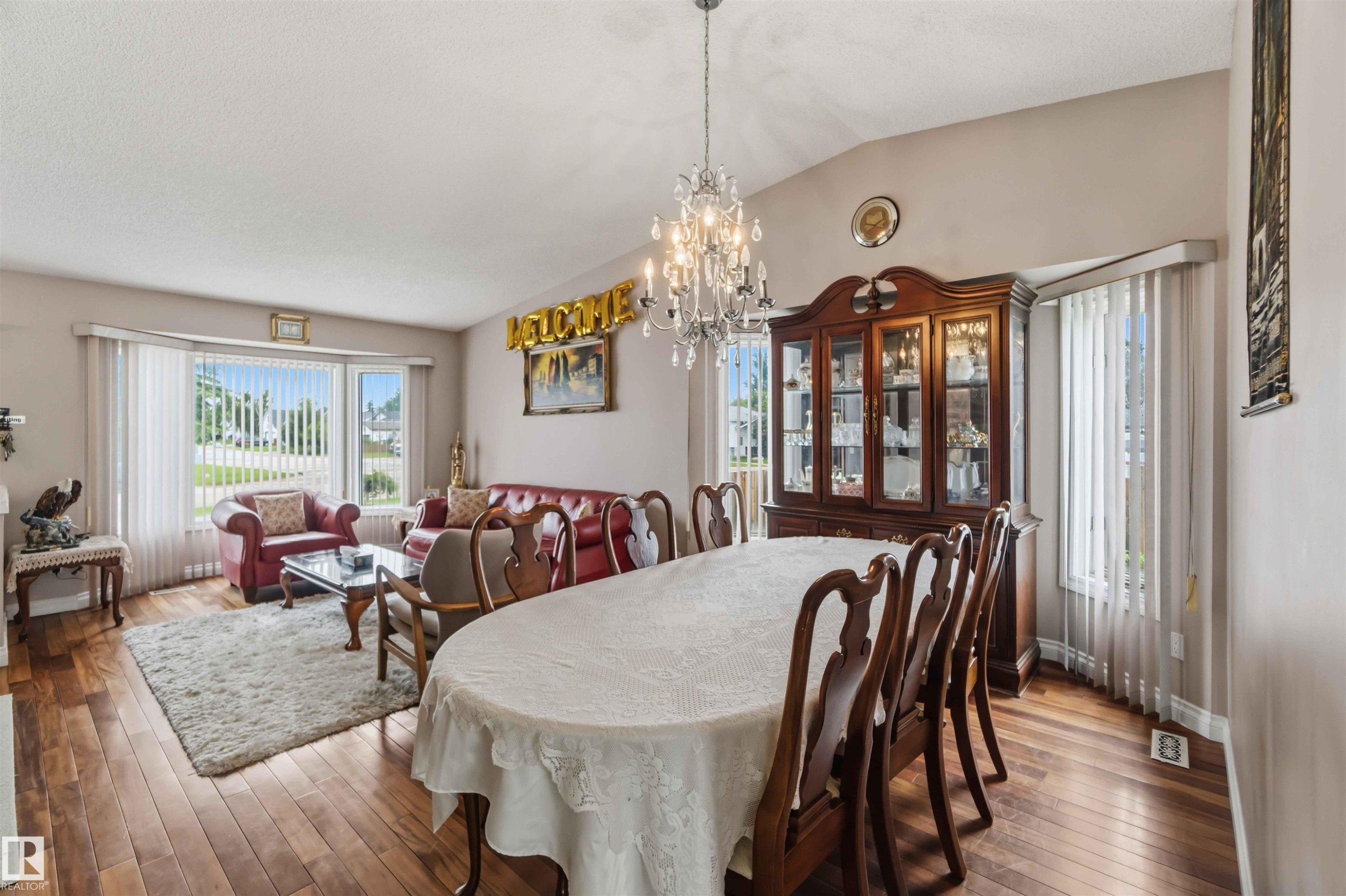 Open-concept living and dining areas featuring wood-finish flooring, a vaulted ceiling with a crystal chandelier, and large windows with vertical blinds - 4428 11A Avenue Nw, Edmonton, AB - Indoor Photo Showing Dining Room