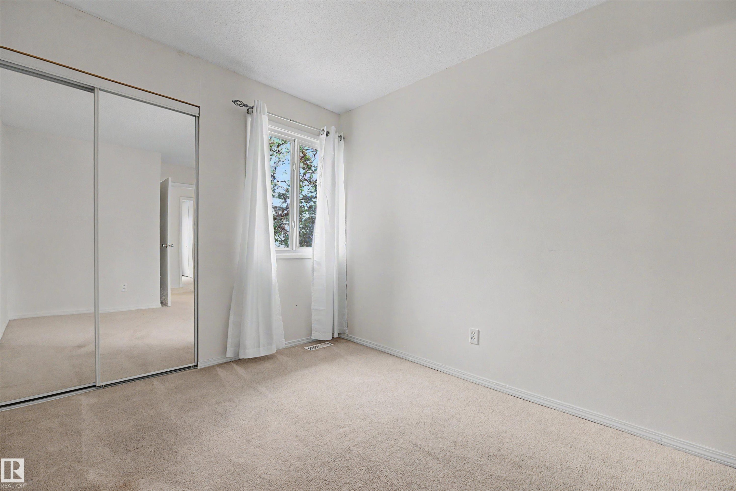 Room featuring neutral-toned carpeting, a window with white curtains, white walls, and a mirrored closet with sliding doors - 198 11421 34 Street, Edmonton, AB