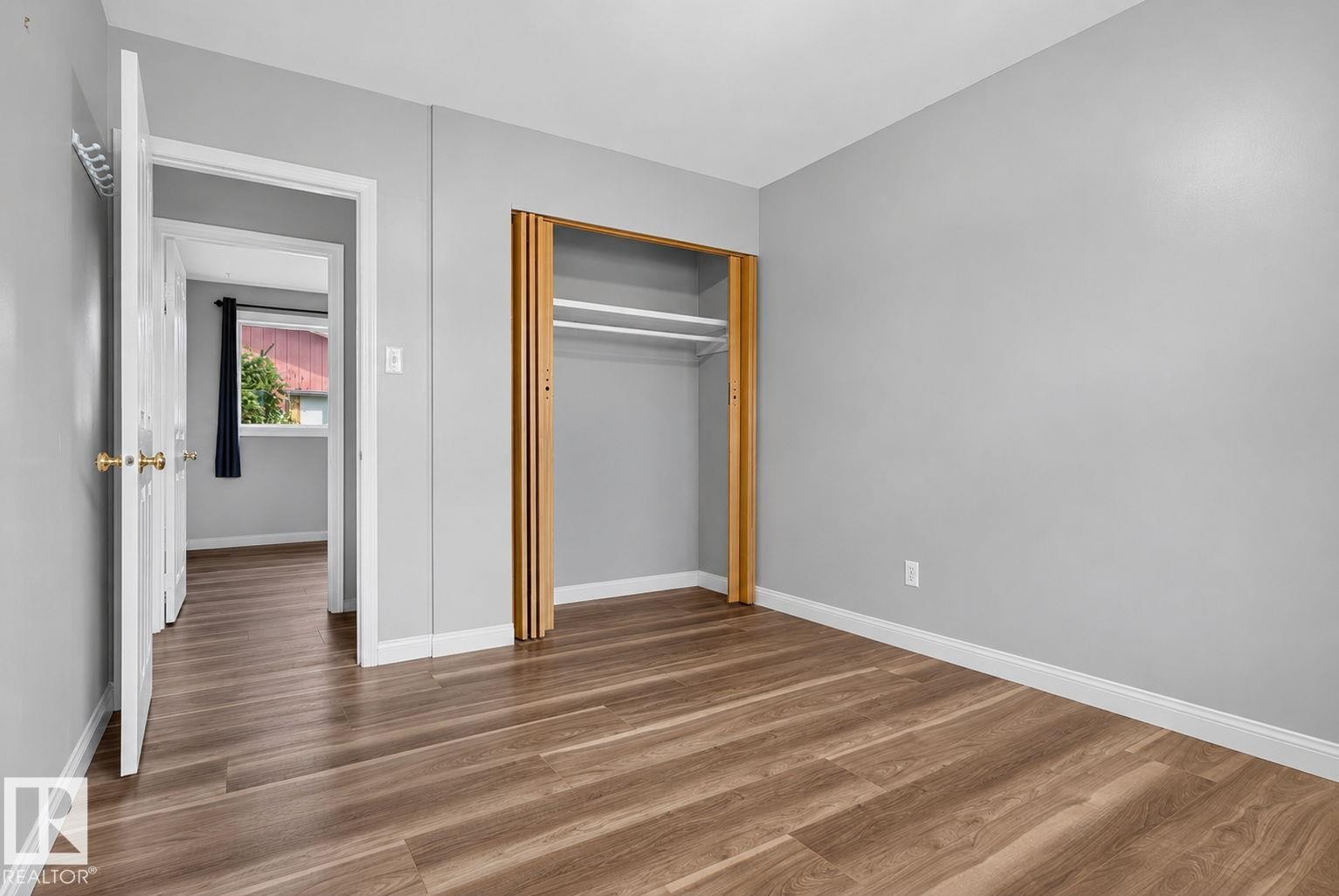 Bedroom featuring wood-finish flooring, light grey walls, white trim, and an open closet with bi-fold doors - 13431 109 Street, Edmonton, AB