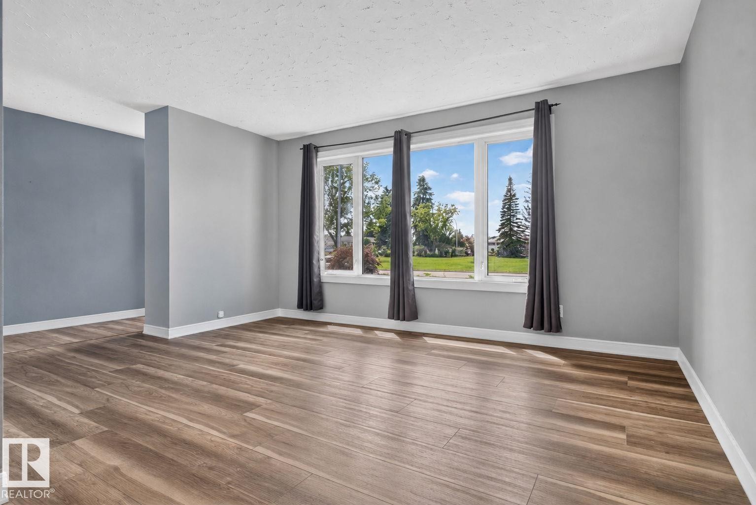 Spacious room featuring wood-finish flooring, a large multi-pane window, and light grey walls with white trim - 13431 109 Street, Edmonton, AB