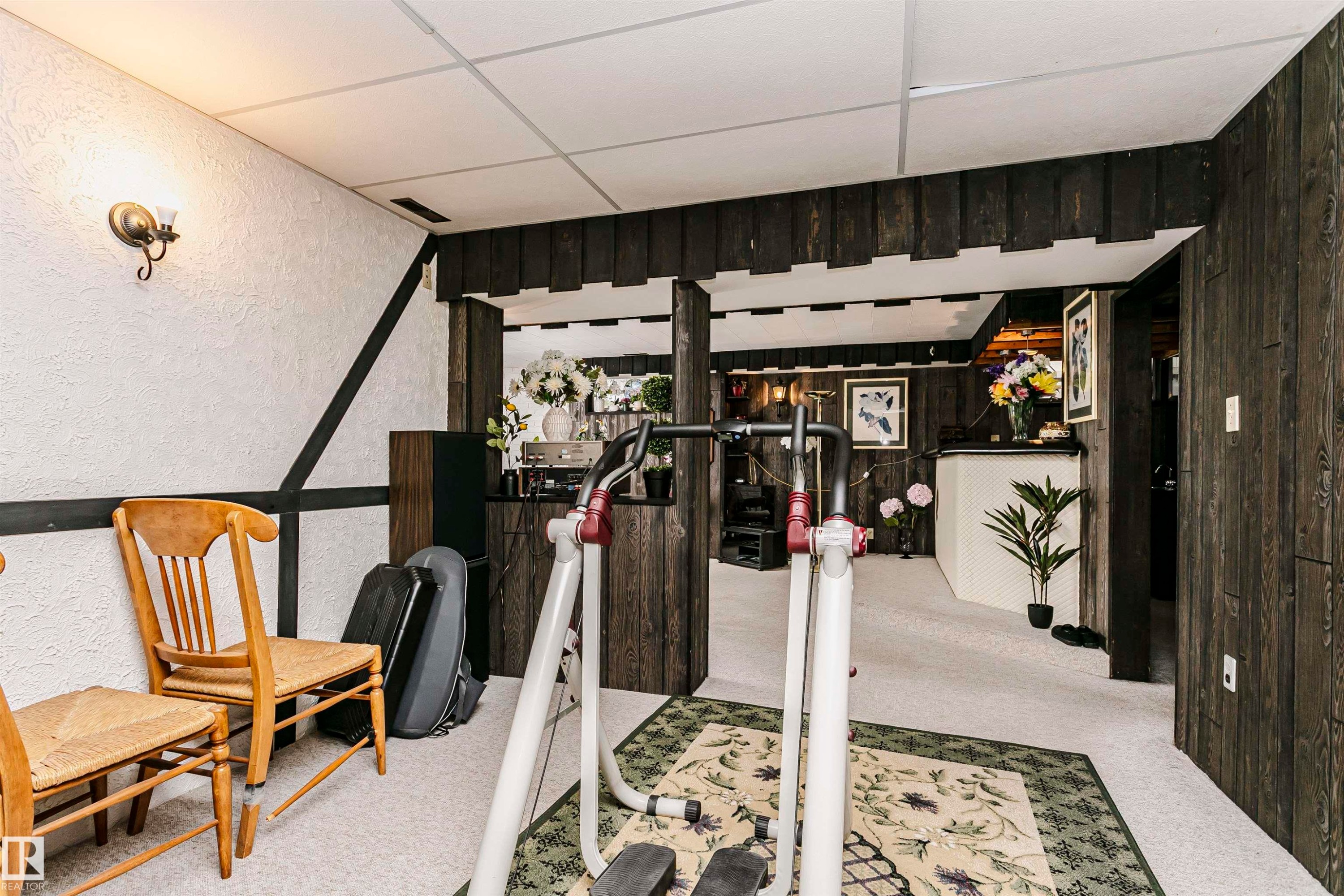 Finished basement space featuring textured white walls with dark trim accents, dark wood-finish wall paneling, light-colored carpeting, and a drop ceiling with integrated lighting - 14231 80 Street, Edmonton, AB